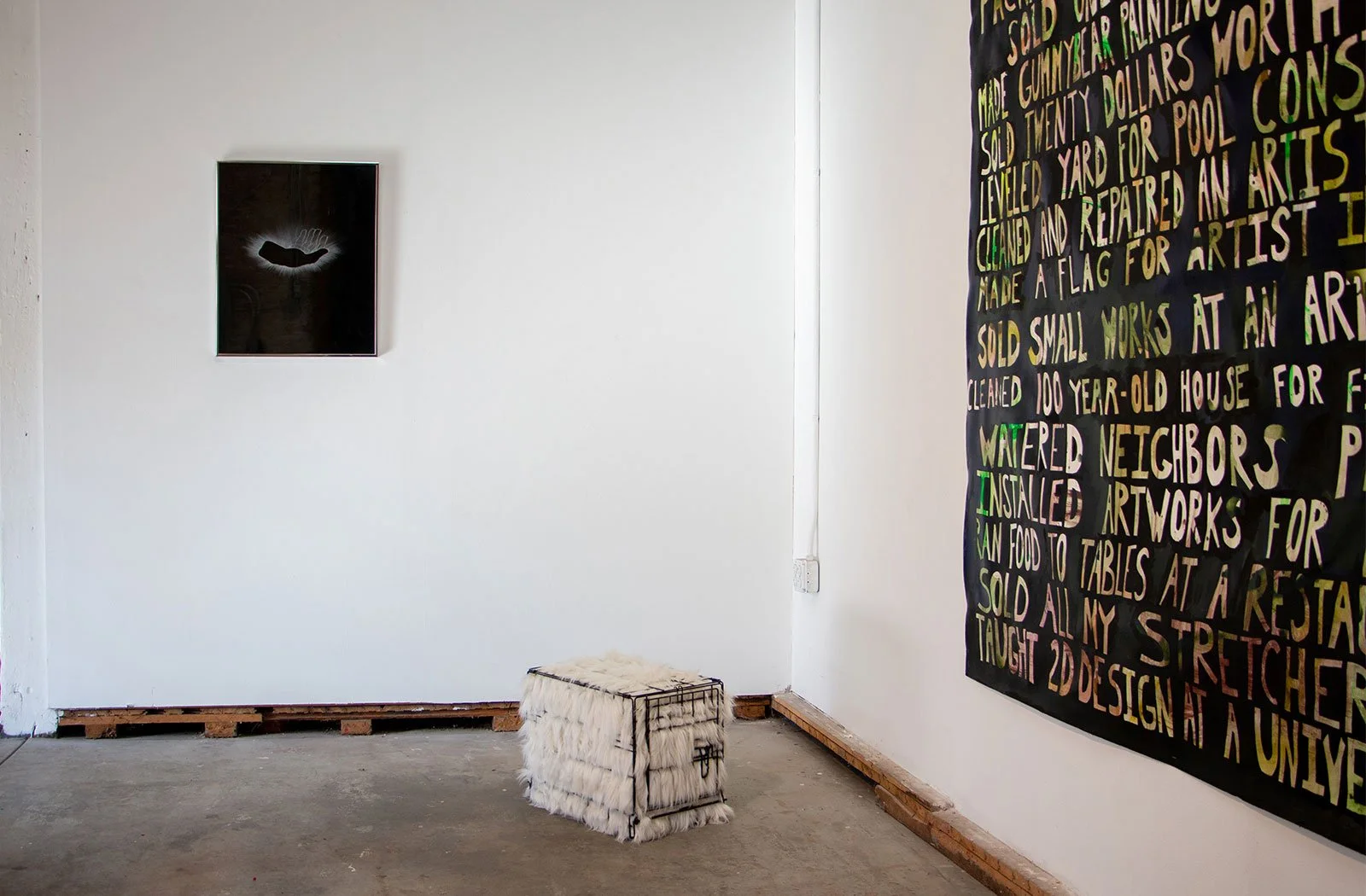An art gallery with a white wall displaying two artworks: a small dark framed piece on the left and a large colorful text-based artwork on the right. There is a fluffy white cube-shaped stool in front of the wall, and the gallery has a concrete floor.