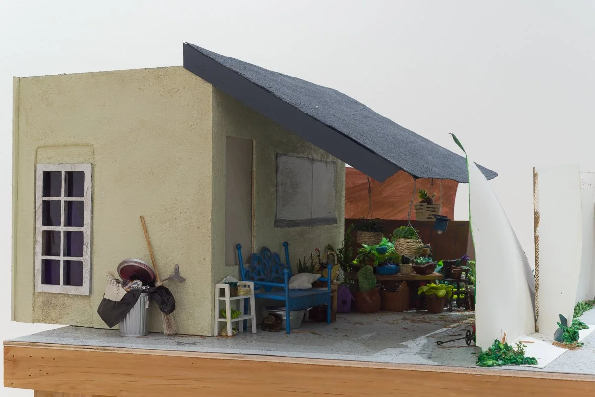 A detail shot of contemporary mixed media sculpture. miniature model of a house with a front porch, a yellow textured wall, a small window, and a blue bench, with a black sloped roof and a white side wall.
