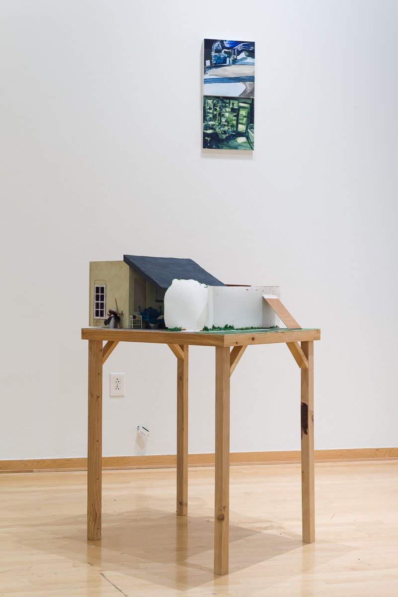 A contemporary art exhibition features Sarah Heyward's sculpture, a miniature architectural model on a wooden table with a white wall background. A painting is hung above the model.