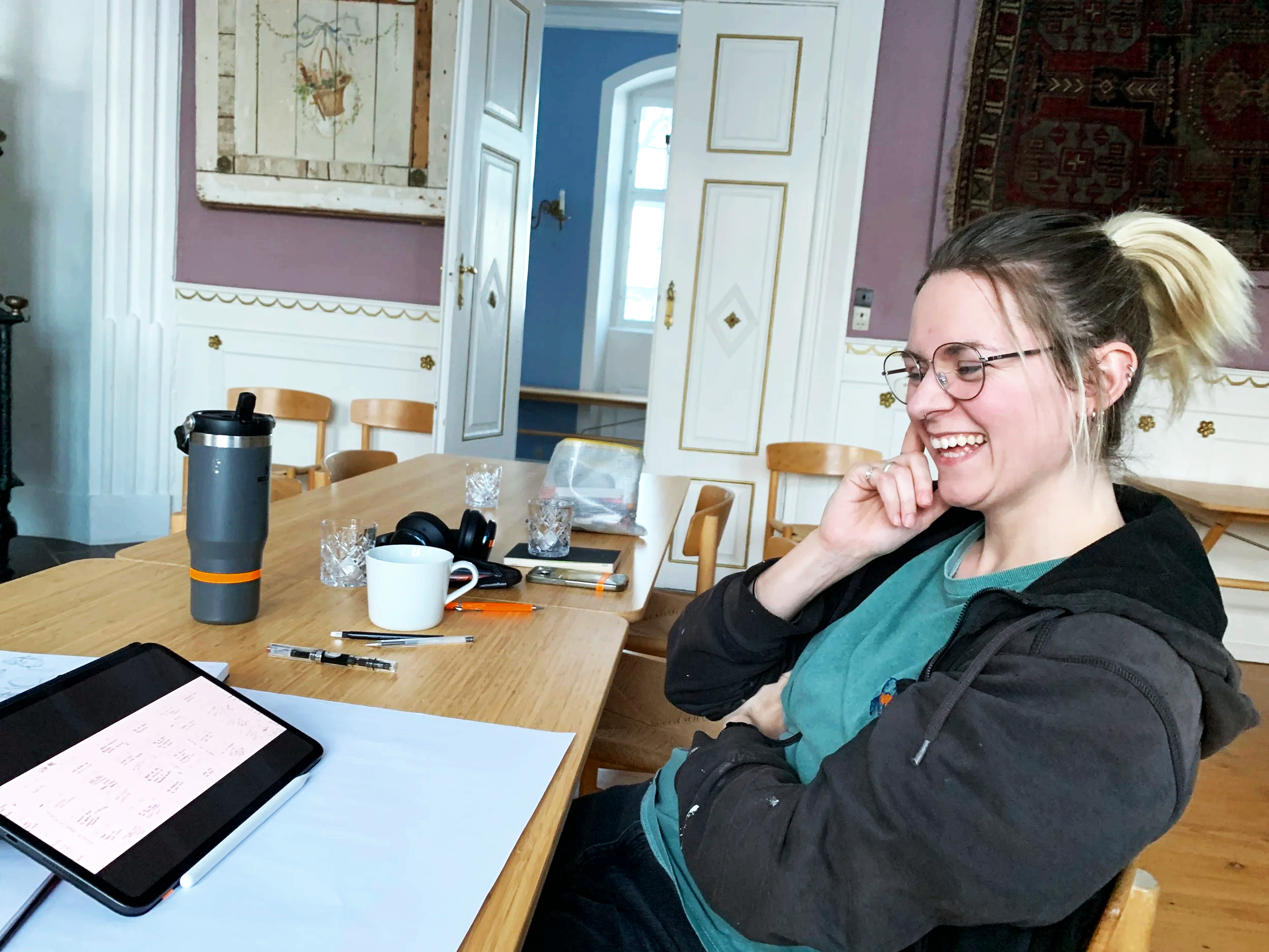 En kvinde med briller smiler og sidder ved et træbord. Der er kopper, en tablet, notesblok og pen på bordet. Bag hende er der et vægmaleri og en åben dør i en klassisk indrettet stue.