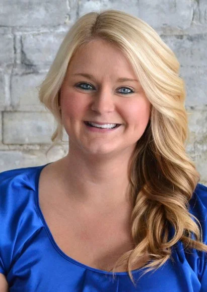 Portrait of a smiling woman with long blonde hair, wearing a shiny blue top, standing against a brick wall.