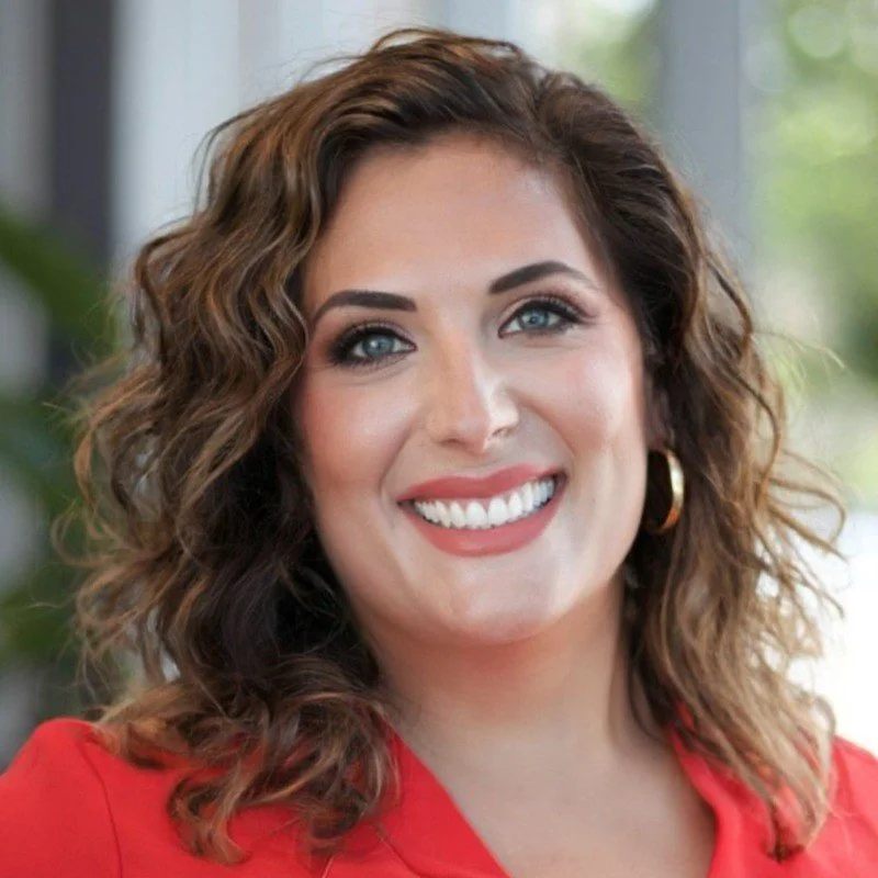 A woman with curly brown hair, blue eyes, and a bright smile, wearing a red top and gold hoop earrings, standing indoors with a blurred background of window and greenery.