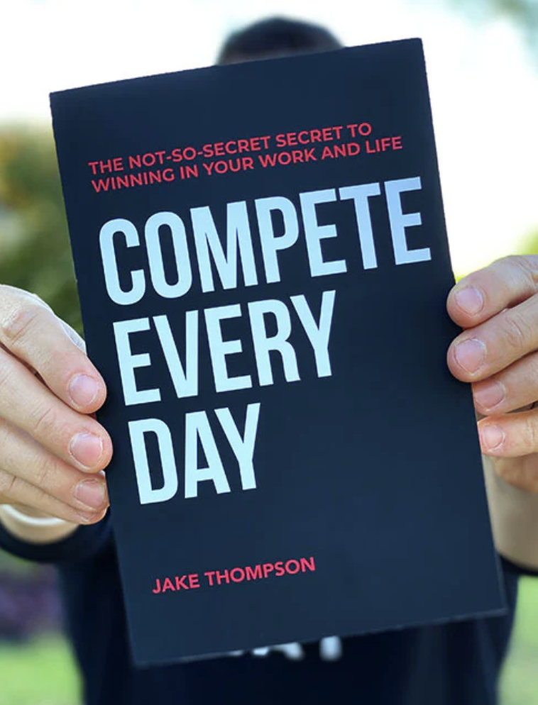 Person holding a book titled 'Compete Every Day' with a subtitle 'The Not-So-Secret Secret to Winning in Your Work and Life' by Jake Thompson.