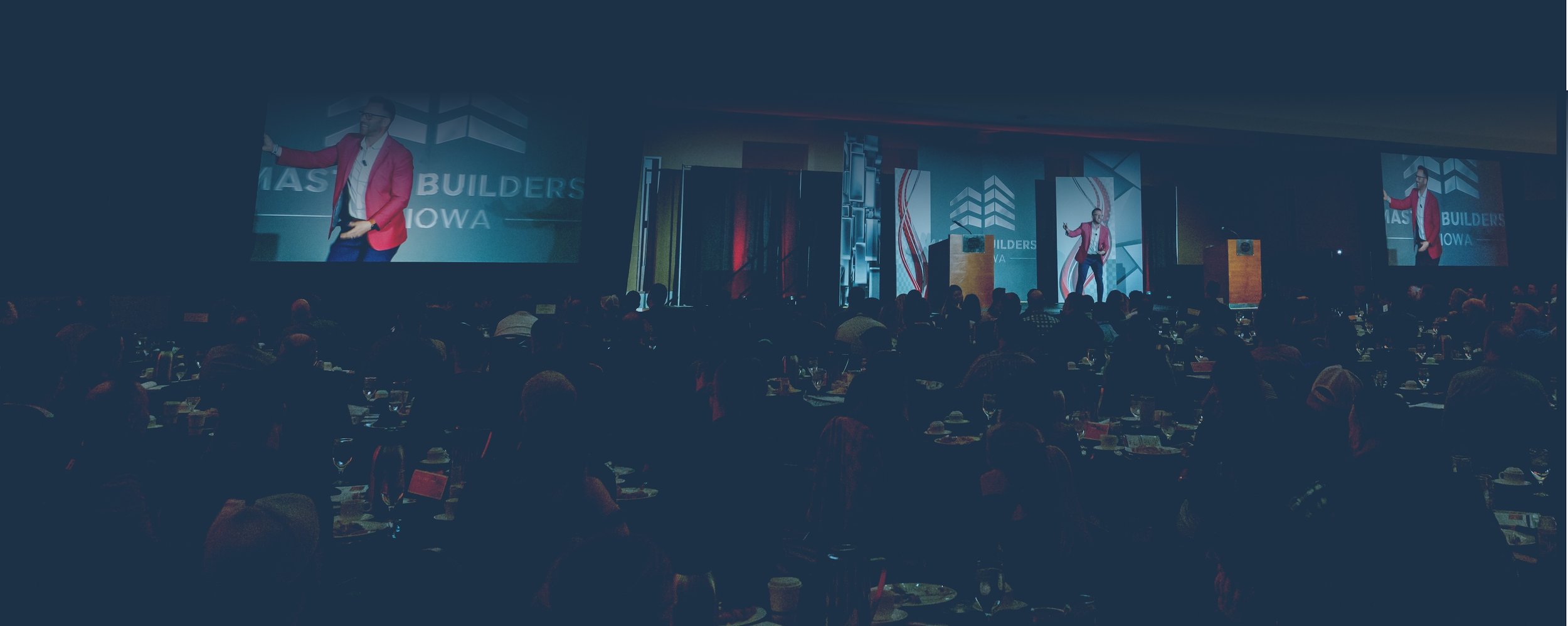 A large conference room filled with attendees seated at tables, watching a speaker on stage and a large screen displaying his image. The event appears to be a professional or business conference.