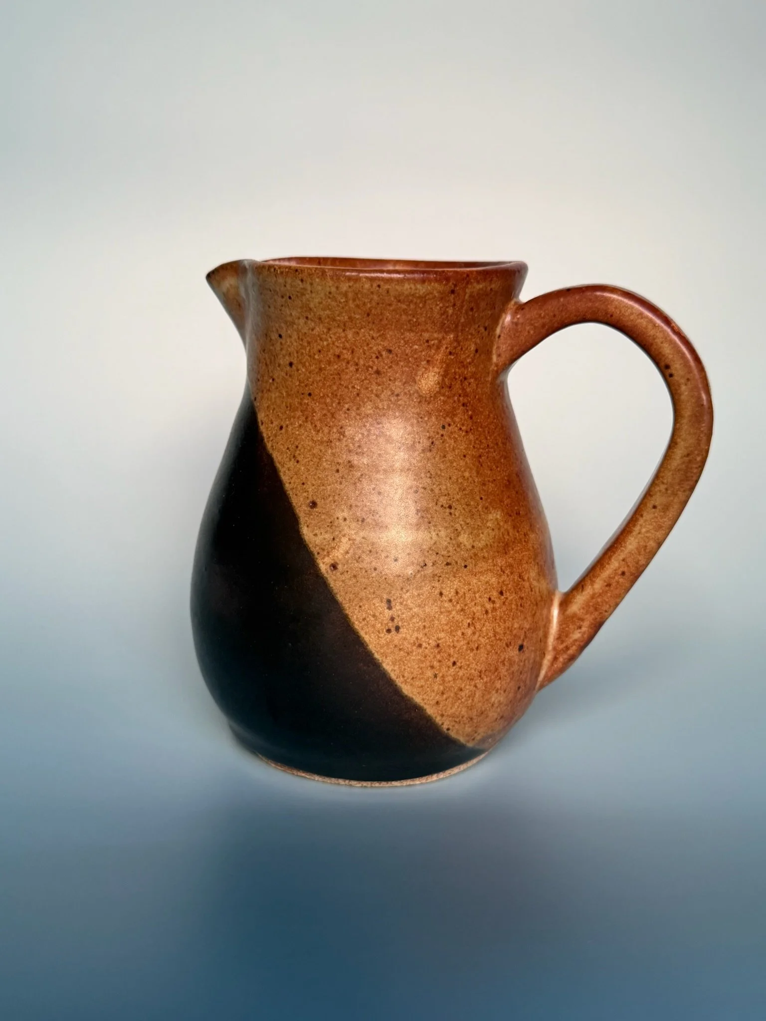 A ceramic pitcher with a gradient of brown and black colors, set against a plain, light background.