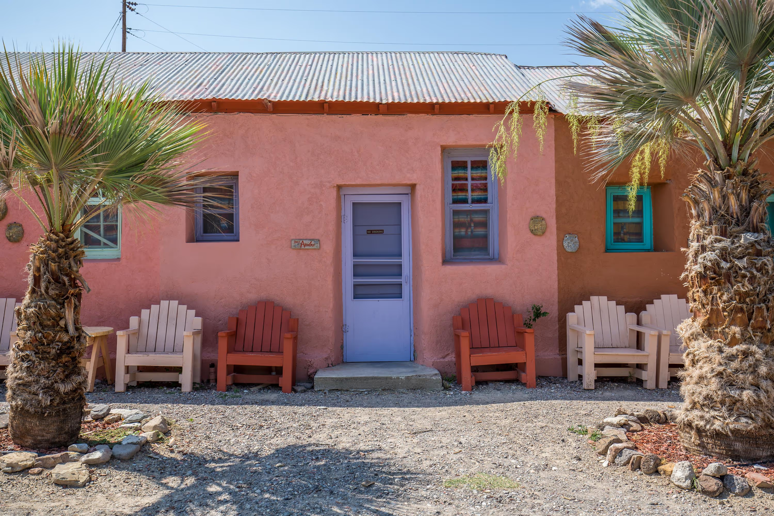 A pink stucco house with a corrugated metal roof, two small windows, and a door in the center. There are white and orange Adirondack chairs outside, flanked by palm trees. A gravel ground with rocks is in the foreground.