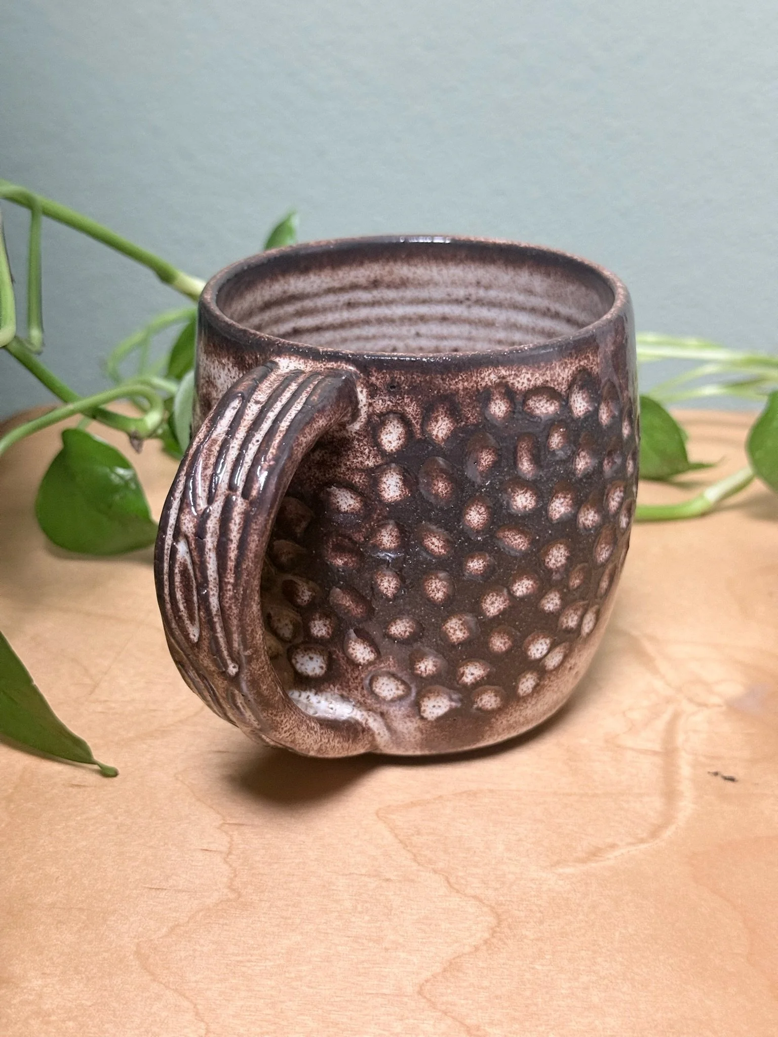 A ceramic mug with a textured, dotted design in brown and cream colors sitting on a wooden surface, with green leafy plant in the background.