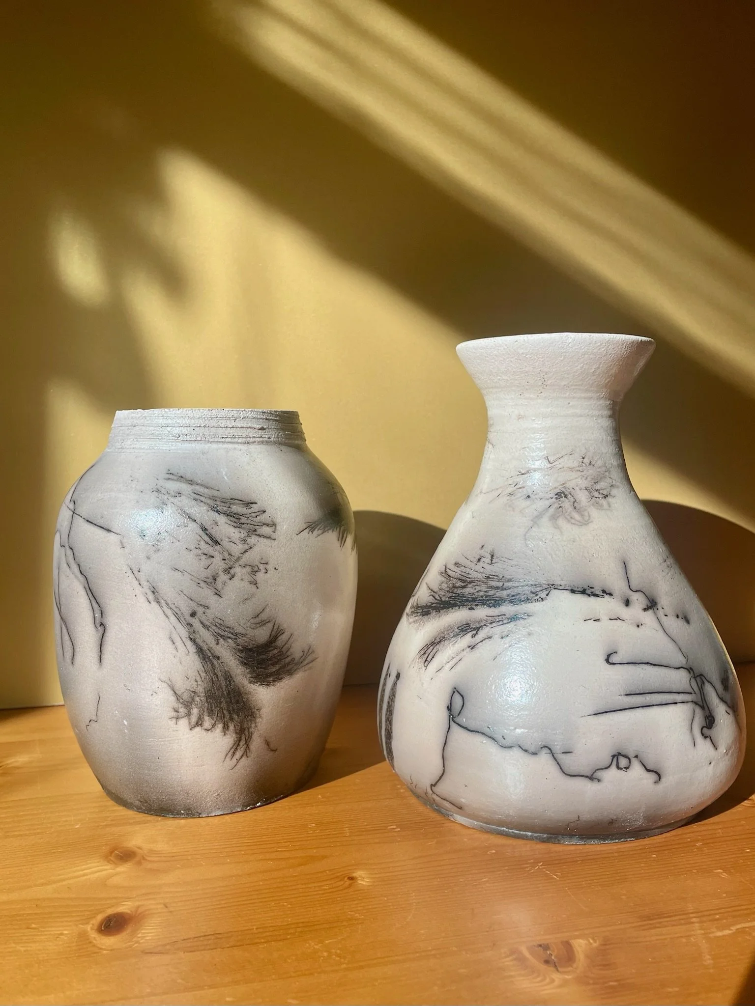 Two white ceramic vases with black abstract line drawings, placed on a wooden surface with a yellow background.