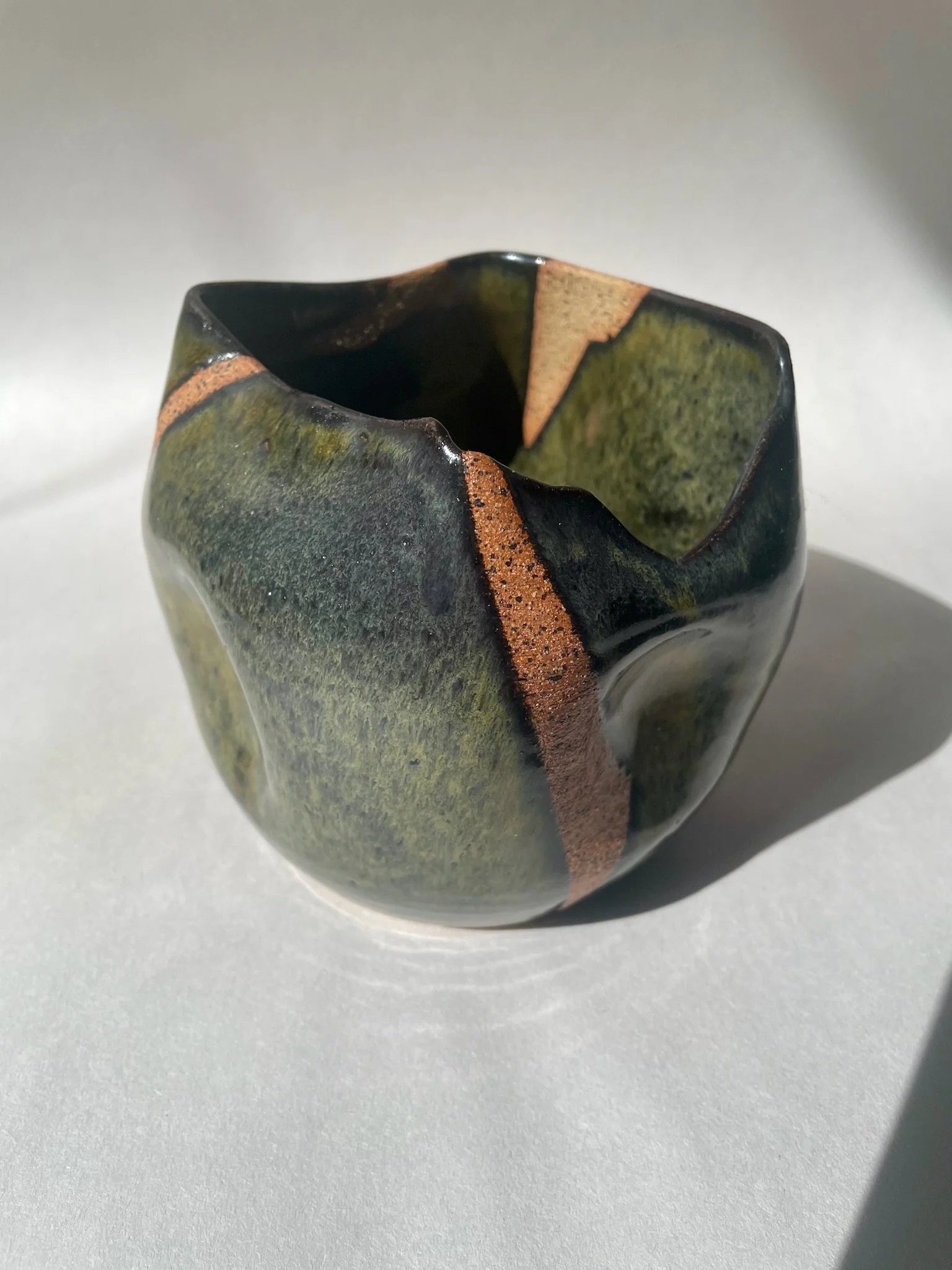 Close-up of a handmade ceramic cup with a wavy rim, glazed in green, black, and earthy tones, on a white surface.