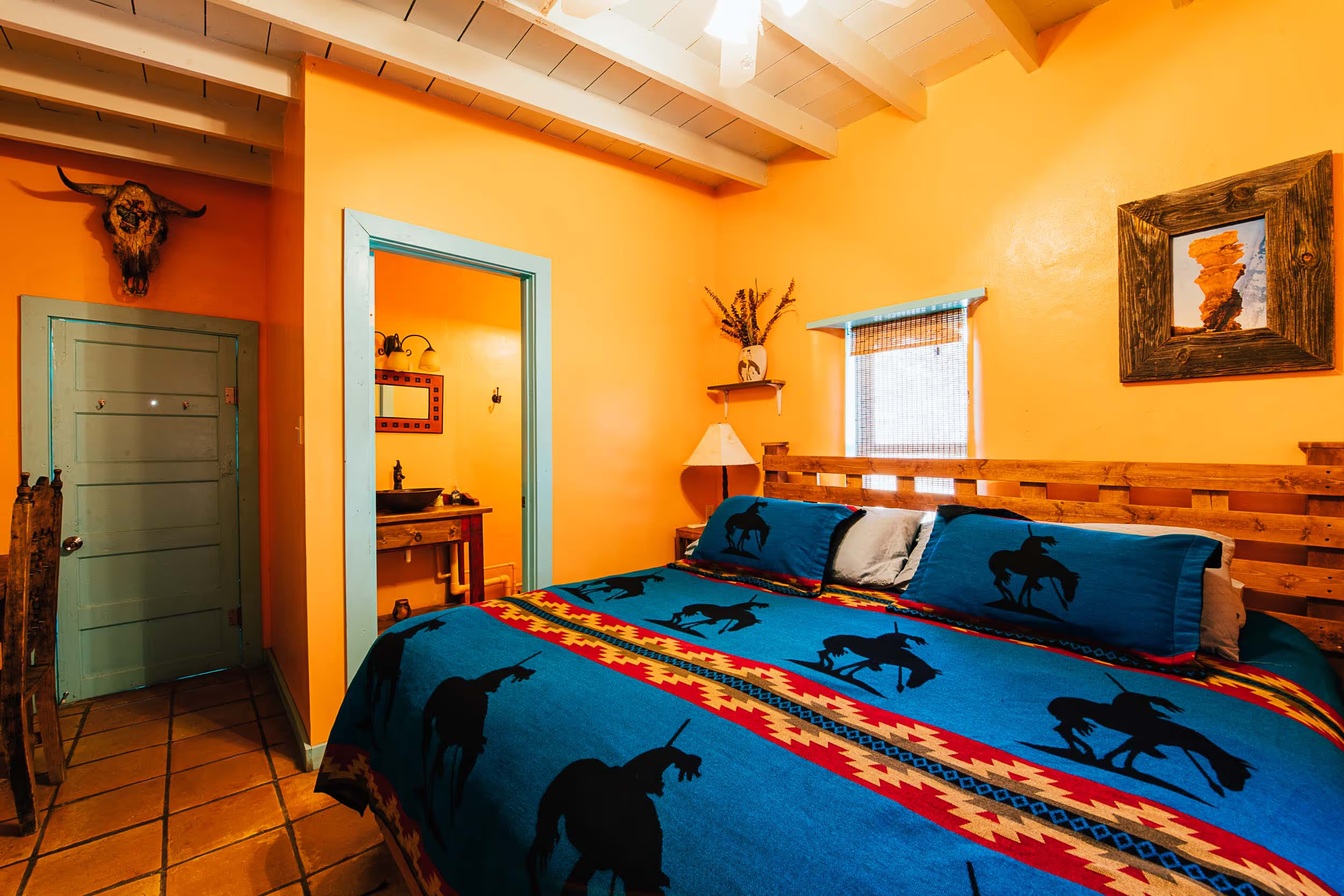A cozy bedroom with bright yellow walls, a wooden bed with a blue bedspread featuring black horse silhouettes, a wooden headboard, and a window with a woven shade. Decor includes a cow skull mounted on the wall, a framed painting, a small shelf with a vase, and a lamp.