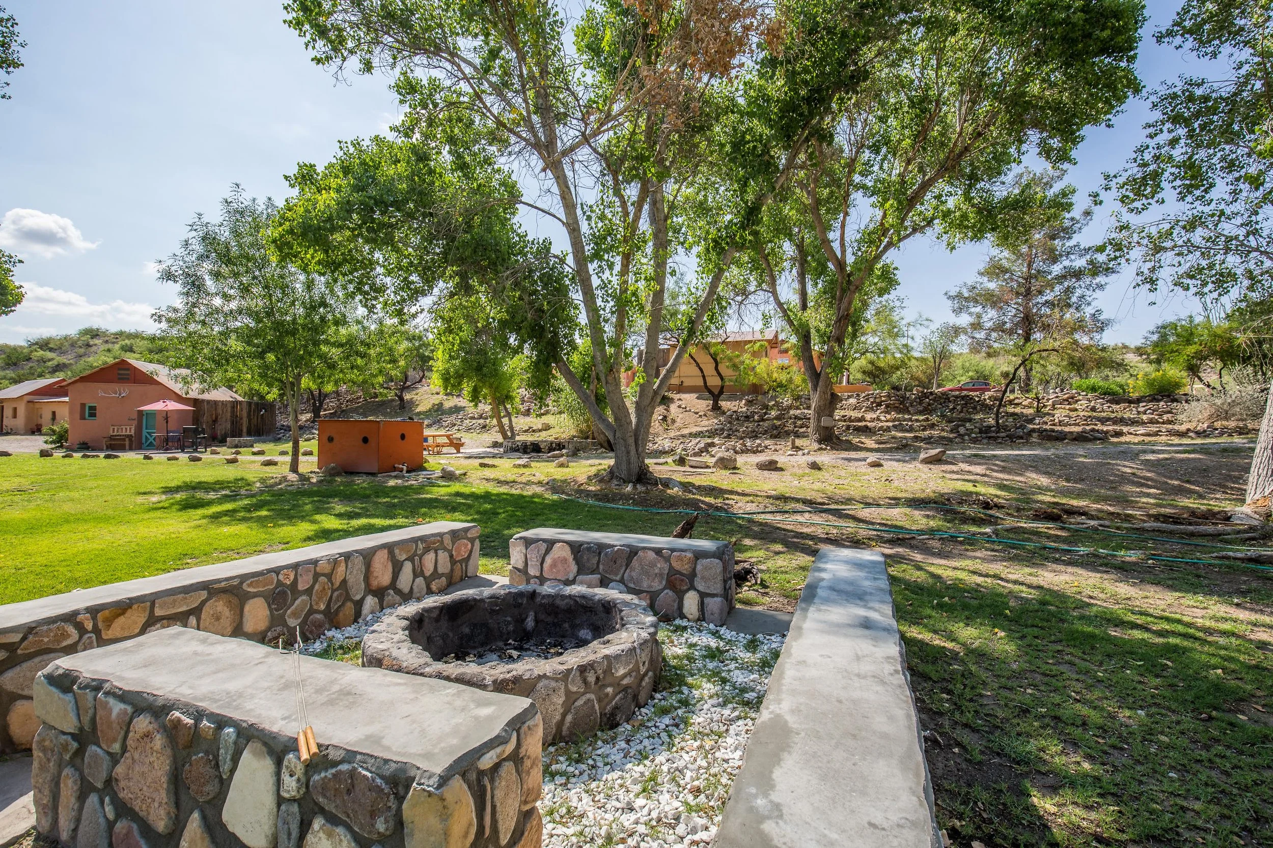 A backyard garden with stone structures, a fire pit, trees, a small house, picnic tables, and a clear sky.