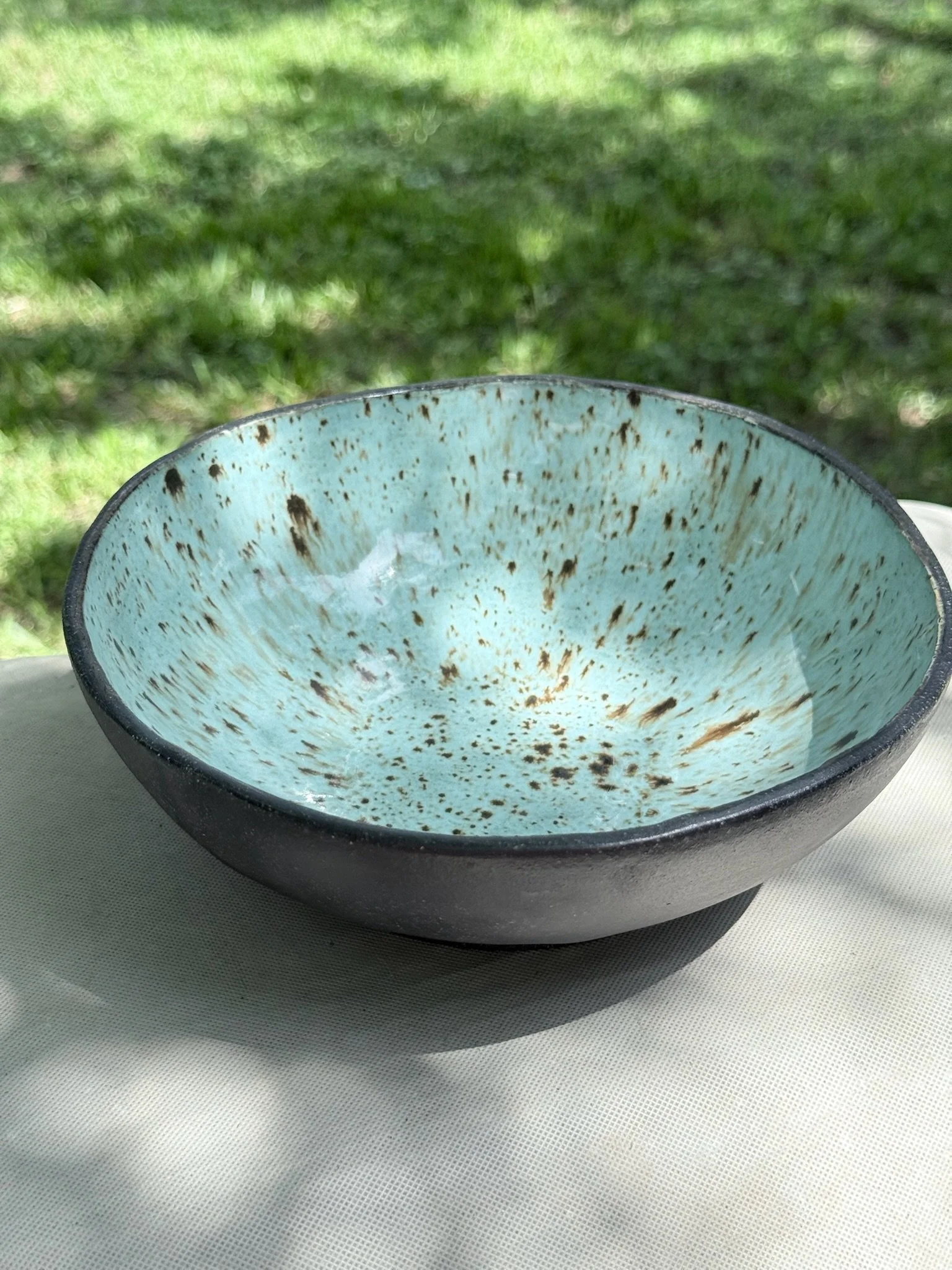 Close-up of a ceramic bowl with a light blue interior and dark speckles, placed on a white textured surface outdoors with grass in the background.