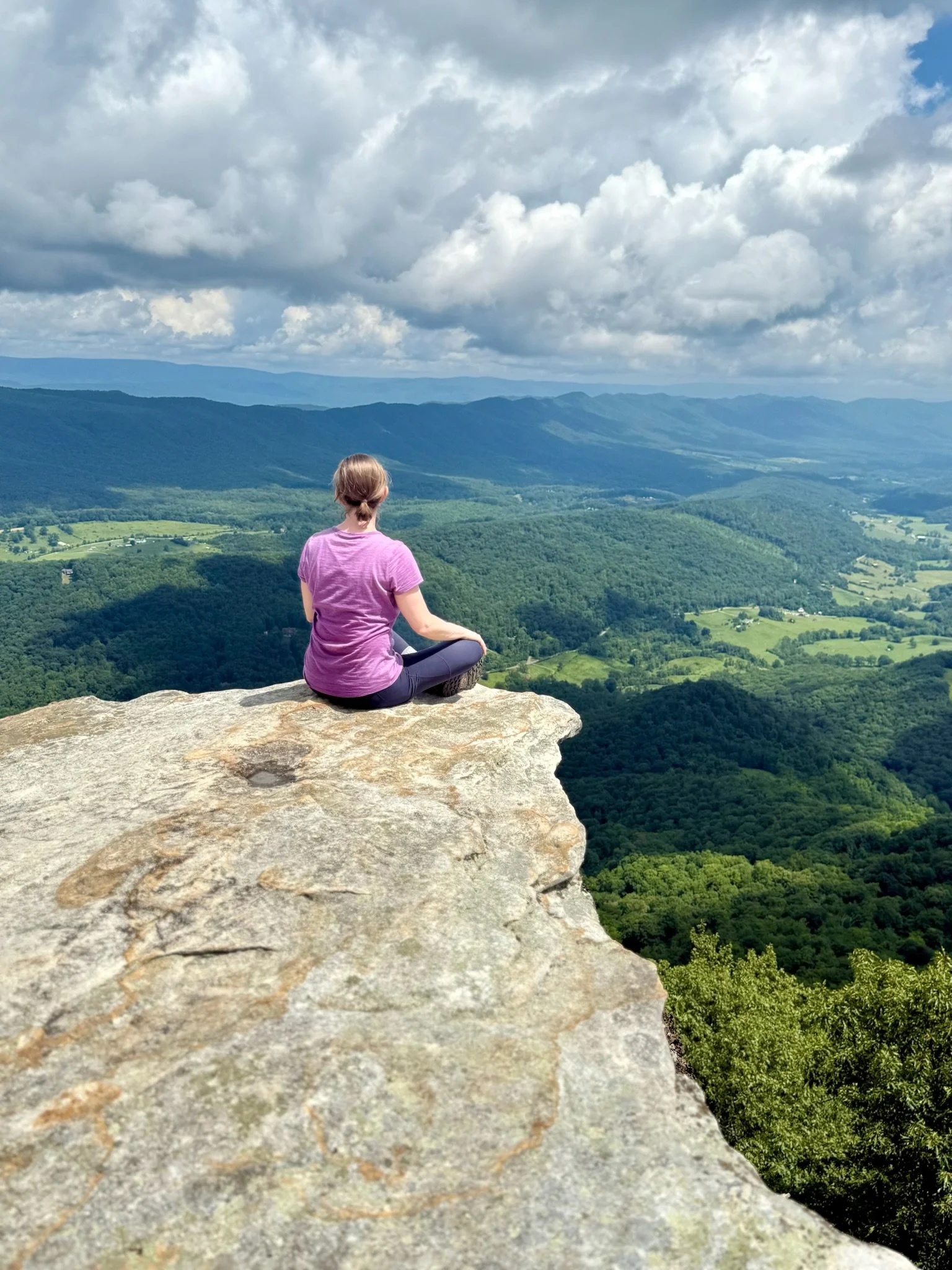 A person sitting on a rocky ledge overlooking a vast green valley and rolling hills, under a sky filled with clouds.