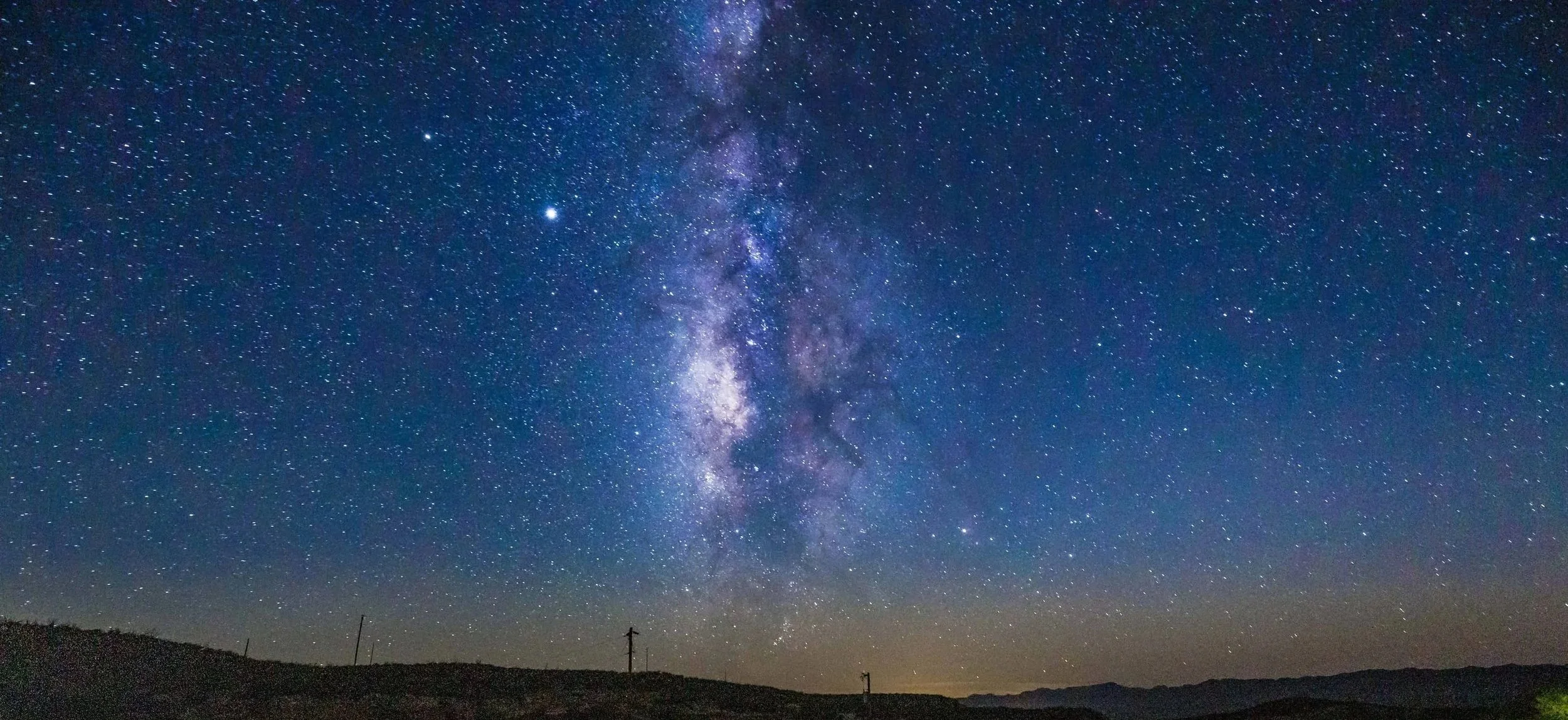 Night sky filled with stars and the Milky Way galaxy visible above a silhouette of a landscape with power lines.