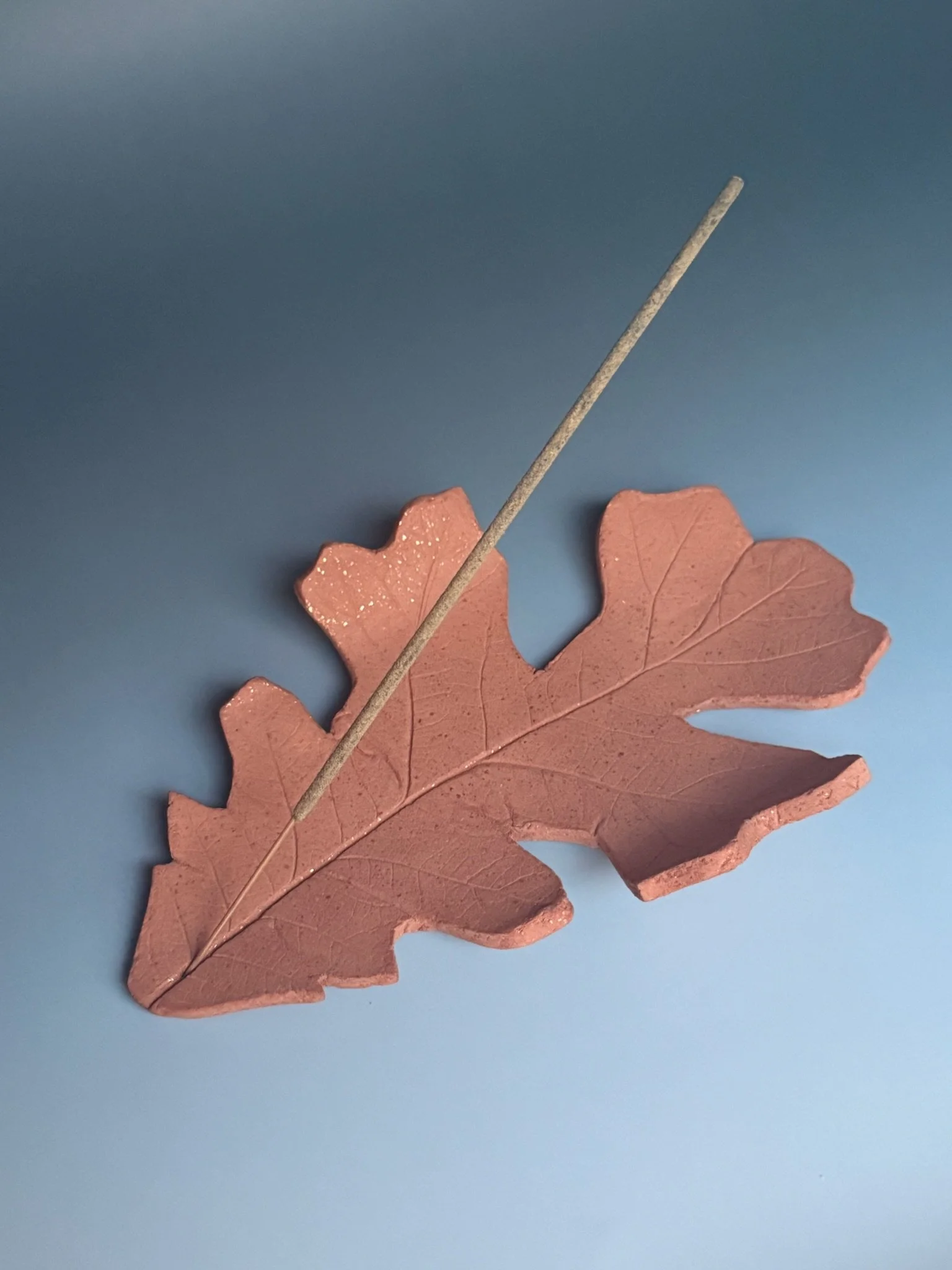 Ceramic oak leaf-shaped dish incense holder with an incense stick, placed on a light blue surface.