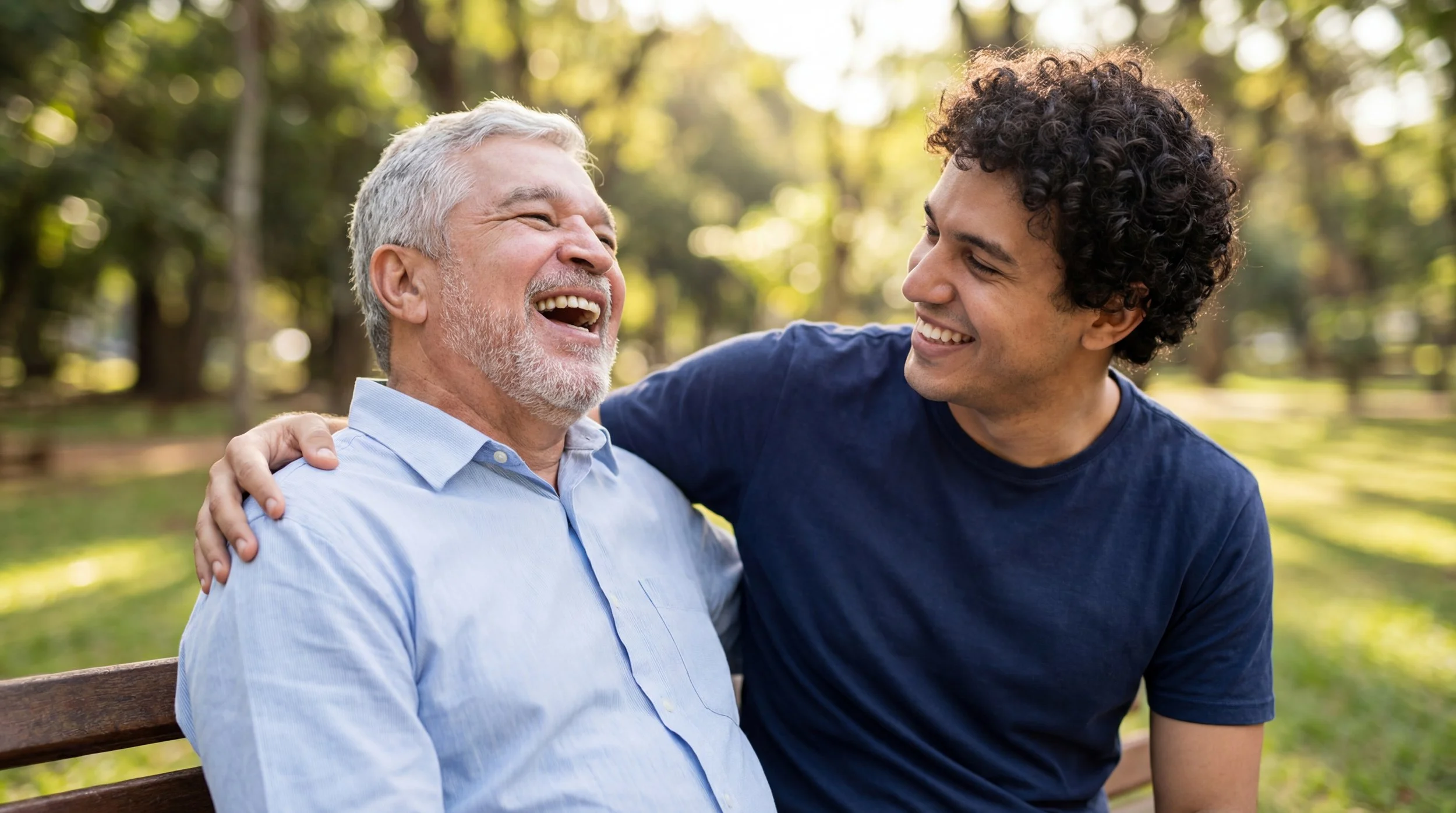 An elderly man laughing and smiling, sitting on a park bench with a young man , both sharing a joyful moment outdoors surrounded by trees and sunlight.