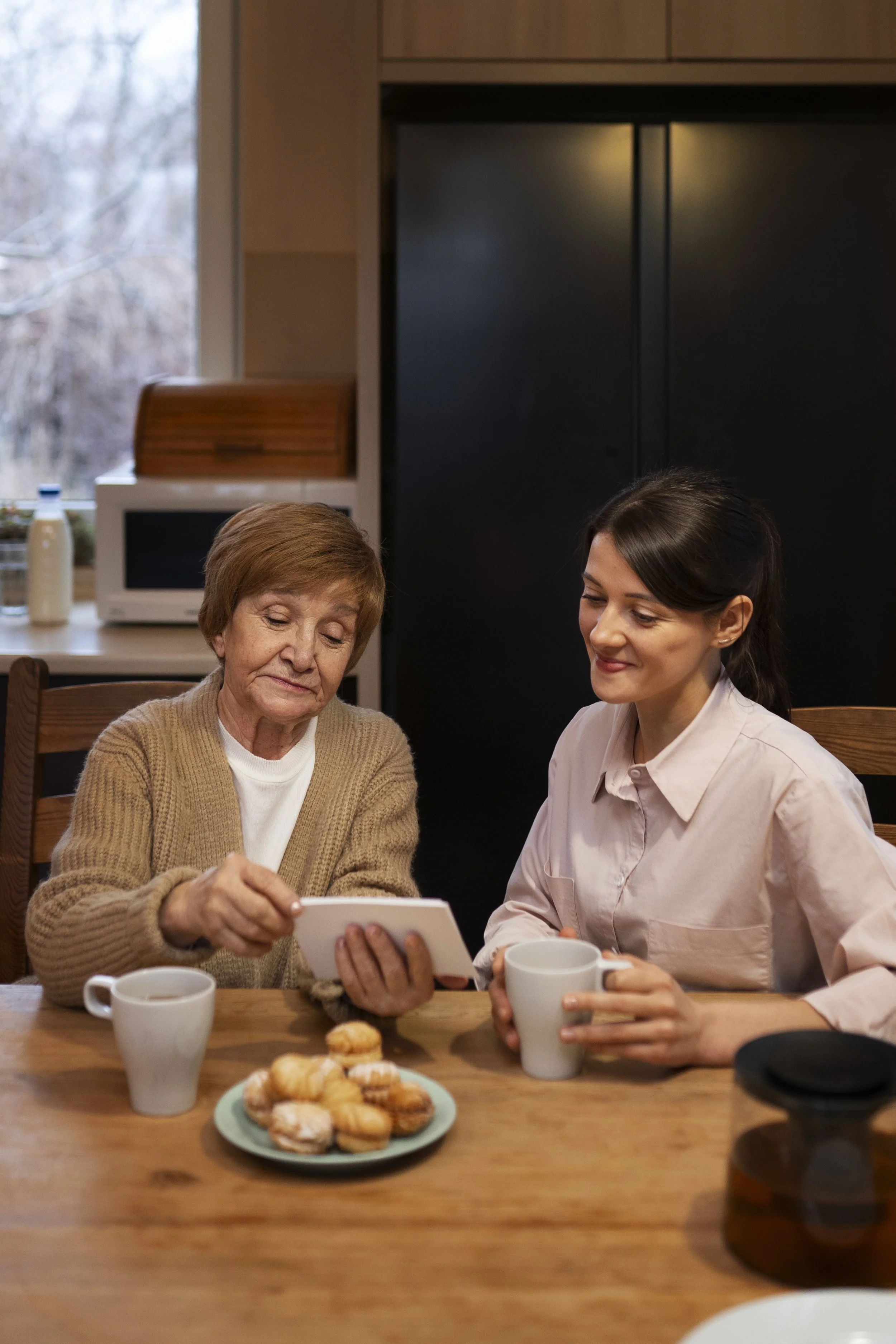 An elderly woman and a young woman sitting at a wooden table in a kitchen, looking at a photo or a device, with mugs and plate of cookies on the table, during daytime.