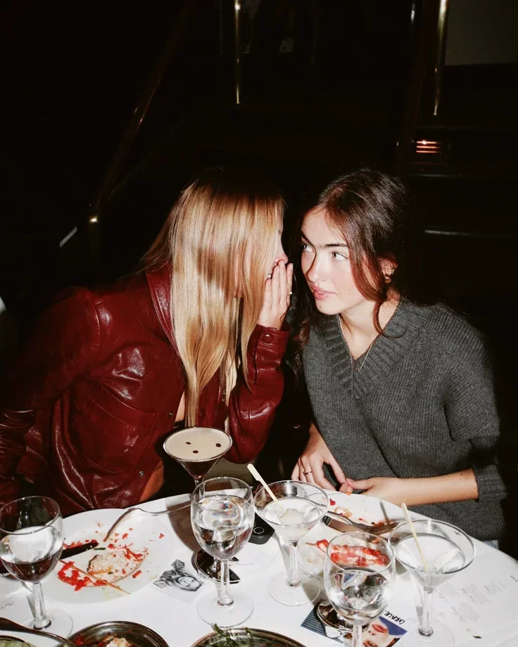 Two women having a secret conversation at a dinner table with drinks and food.