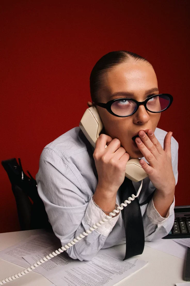 A woman with glasses, dressed in a business shirt and tie, talking on a landline phone with a stressed or exhausted expression, sitting at a desk with papers and a calculator in front of a red background.