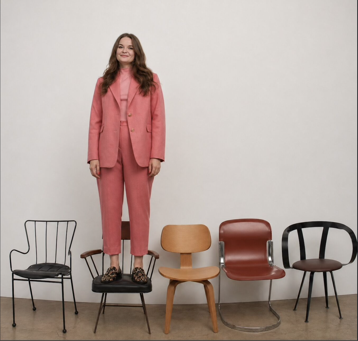 A woman in a pink suit standing on a black chair, with four other chairs of different styles and colors lined up against a plain white wall.