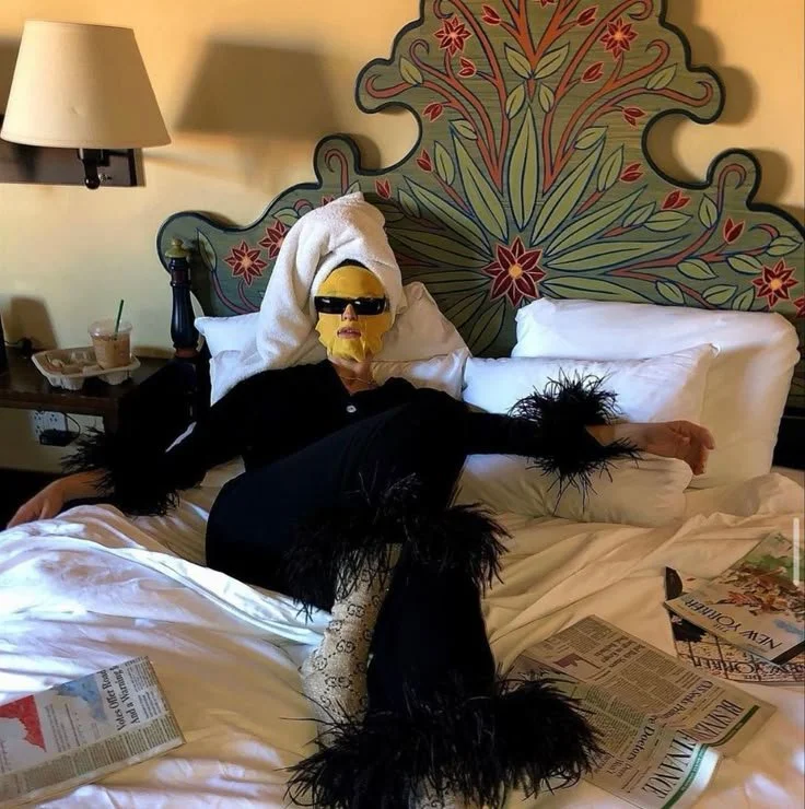 Person lying on a bed with a decorative headboard, wearing sunglasses, a towel wrapped as a hair, a black outfit with feathered sleeves and boots, magazine pages spread on the bed, and a side table with drinks and a lamp.