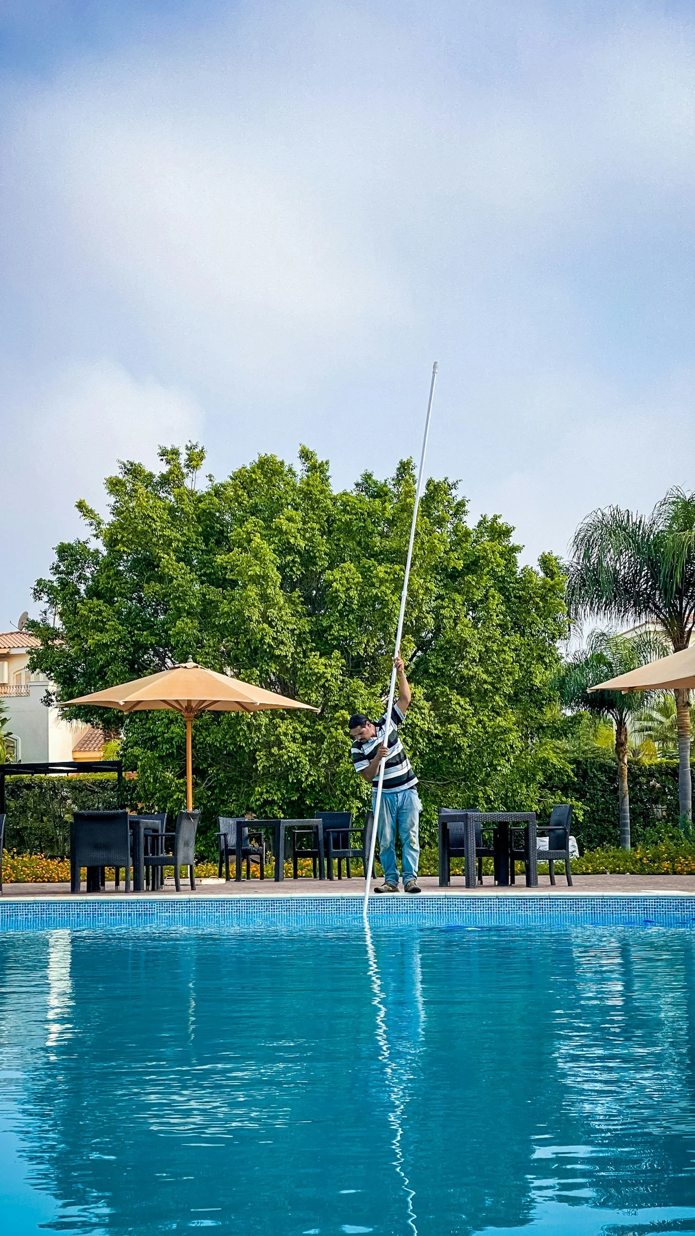 A man by a swimming pool holding a long pole, possibly setting up or adjusting a flag or similar item, with outdoor seating and large umbrellas, green trees, and a partly cloudy sky in the background.