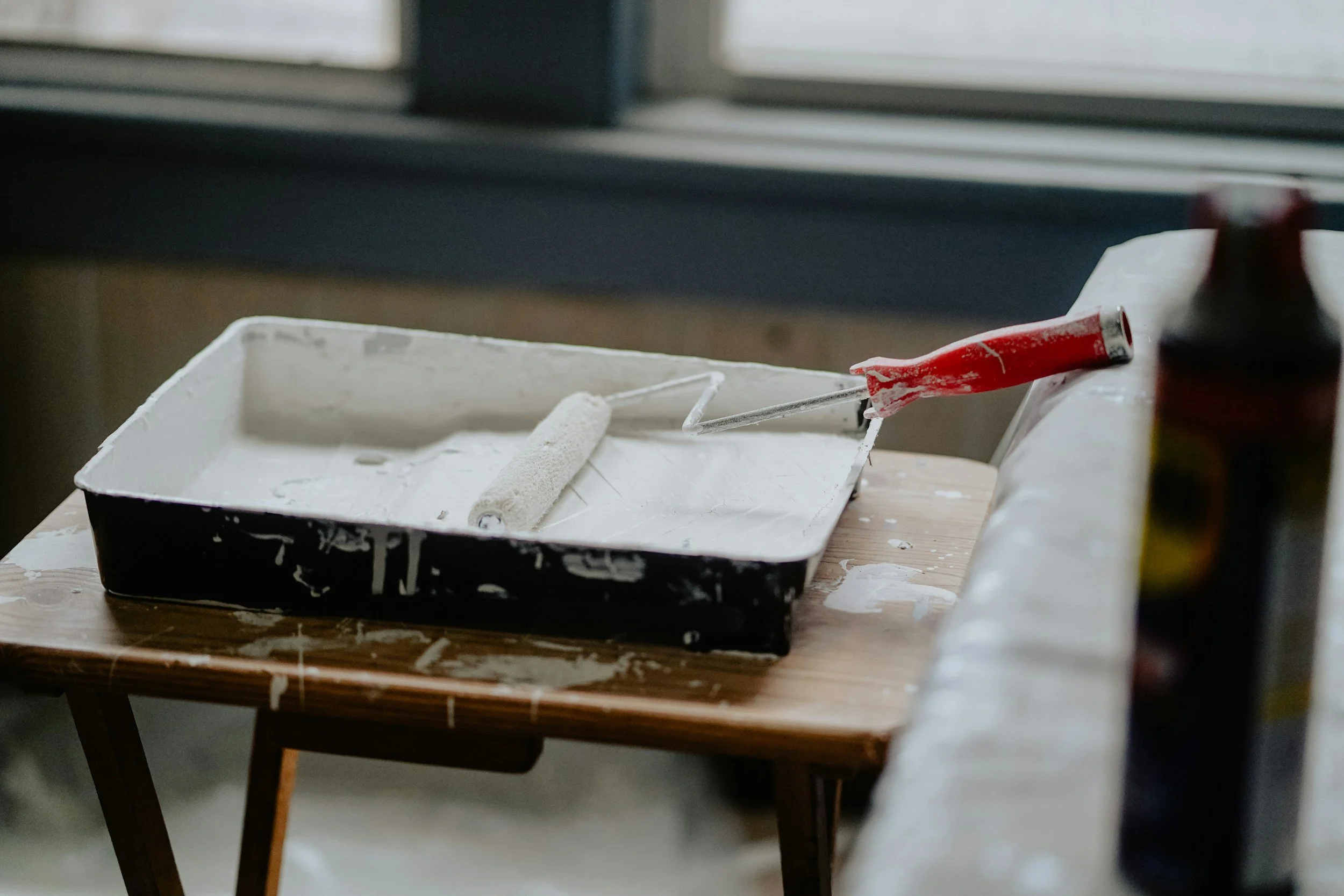 A workbench with a paint roller covered in white paint, a paintbrush, and bottles of paint on a wooden table in front of a window.