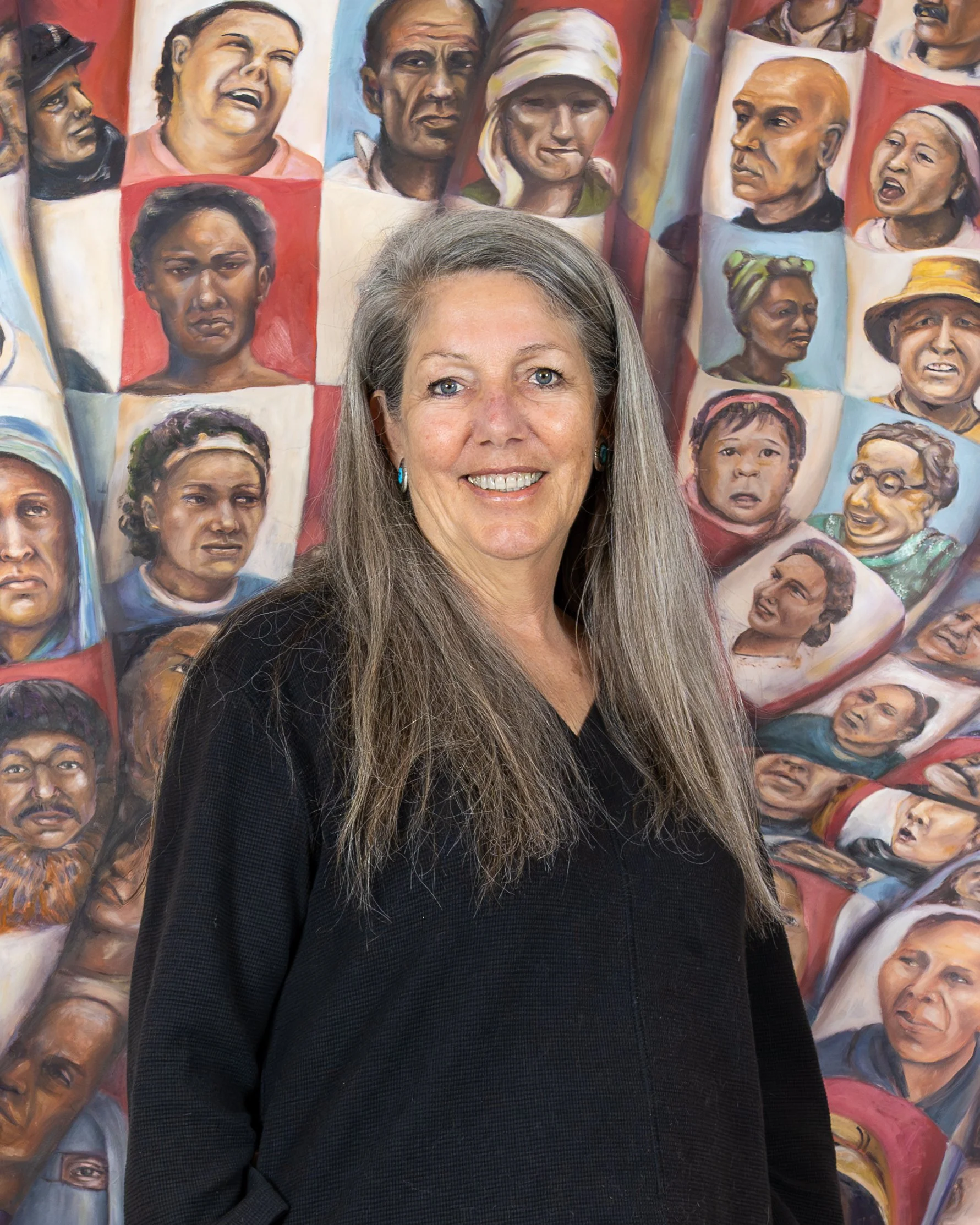 A woman with long gray hair, wearing earrings, smiling, standing in front of a mural of diverse faces painted in a realistic style.