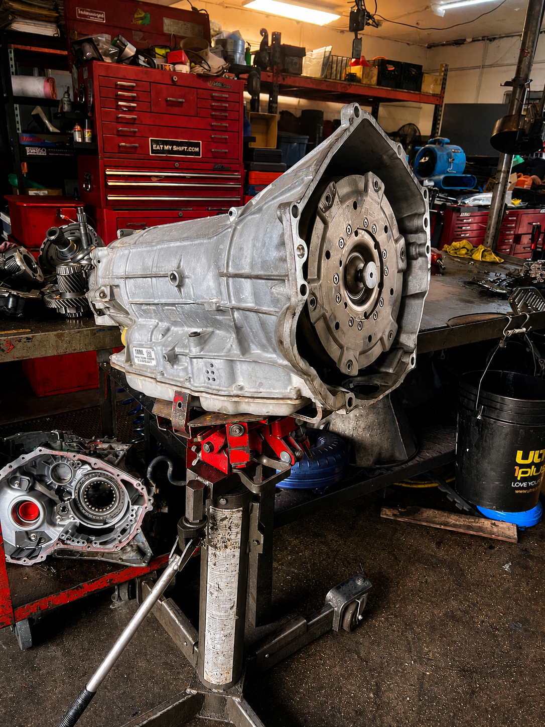 An automotive transmission mounted on a repair stand in a cluttered garage, with tools and spare parts nearby.