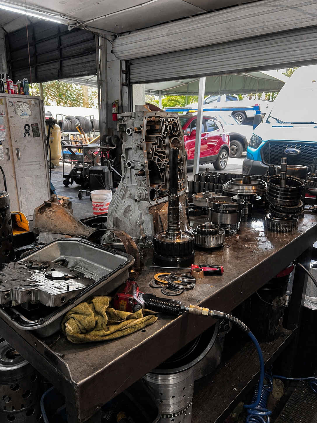 Auto repair shop workbench with disassembled car transmission parts and tools, with cars outside visible through the shop's open garage door.