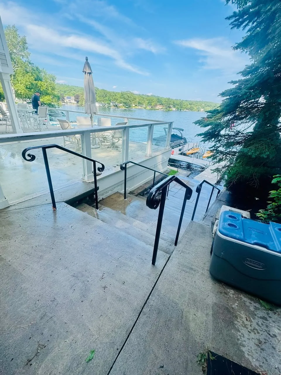 View of a lakeside patio with outdoor furniture, umbrella, boat dock, and a cooler, surrounded by lush trees on a sunny day.