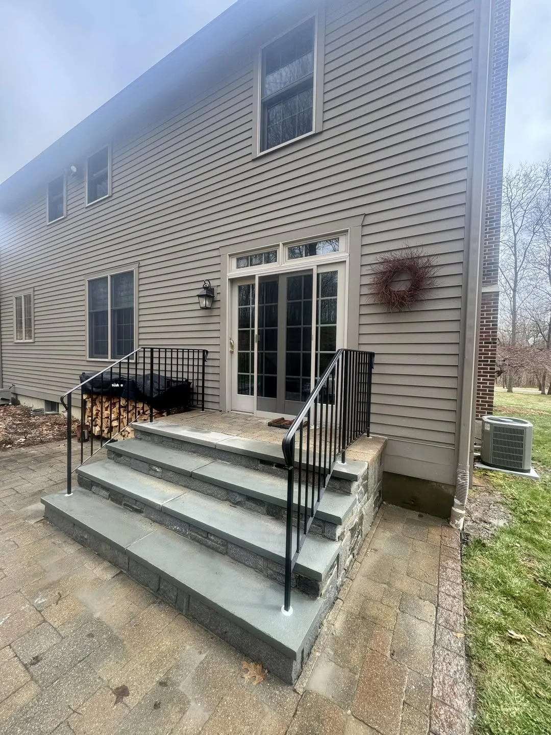 Backyard concrete stairs with black metal railings leading to a sliding glass door on a beige house. The area has a pile of firewood and a wreath on the house wall. The background shows a lawn with trees and an air conditioning unit.