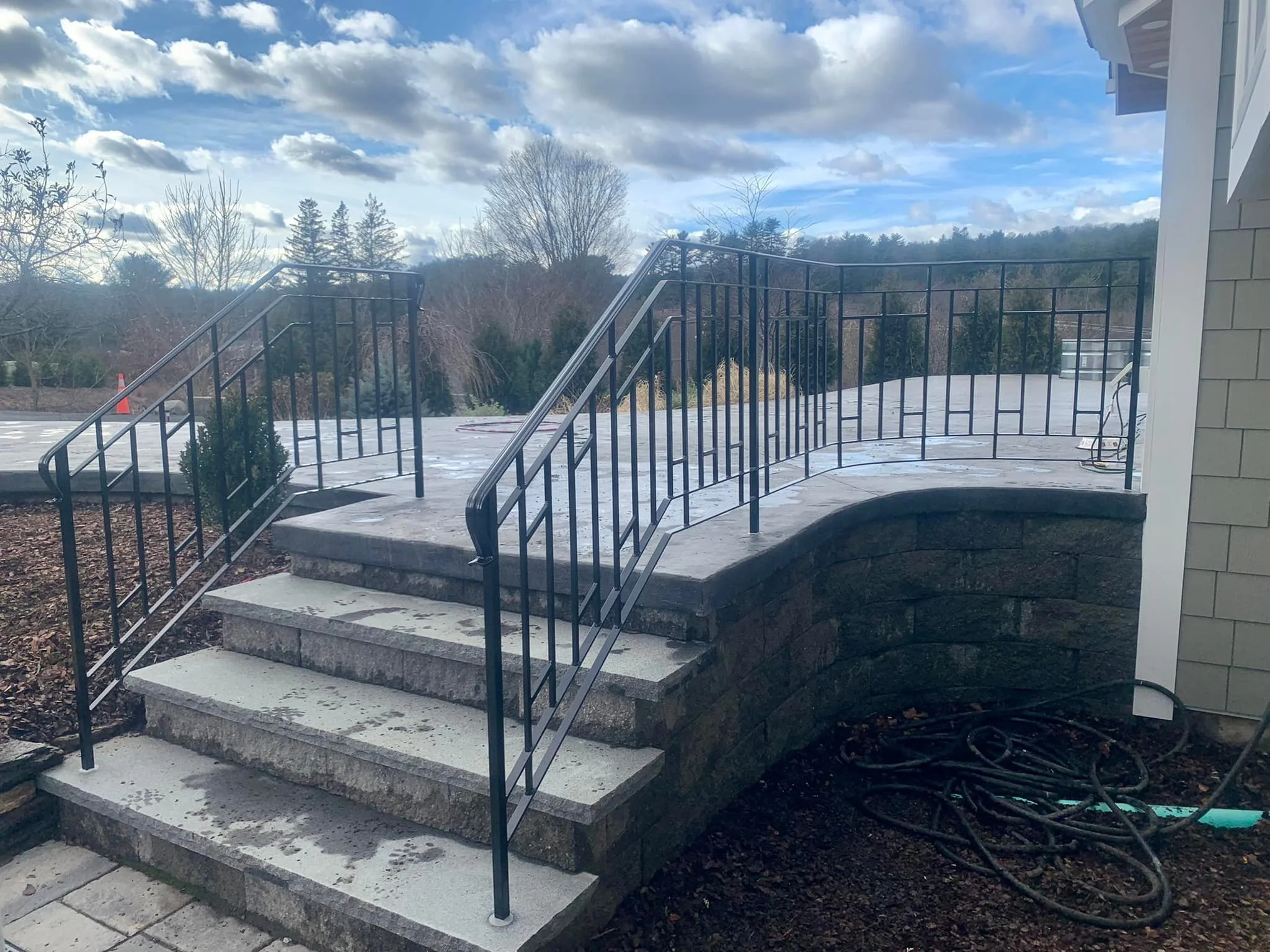 Outdoor stairway with black metal handrails leading up to a concrete platform, with trees and a cloudy blue sky in the background.