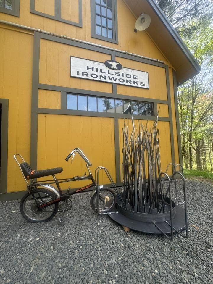 A yellow building with a sign that reads 'Hillside Ironworks'. In front of the building, there is a tricycle and a circular metal rack with tall metal rods.
