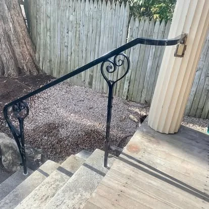 Outdoor staircase with a black metal handrail featuring decorative scrolls, leading down to a gravel area, with a wooden fence and trees in the background.