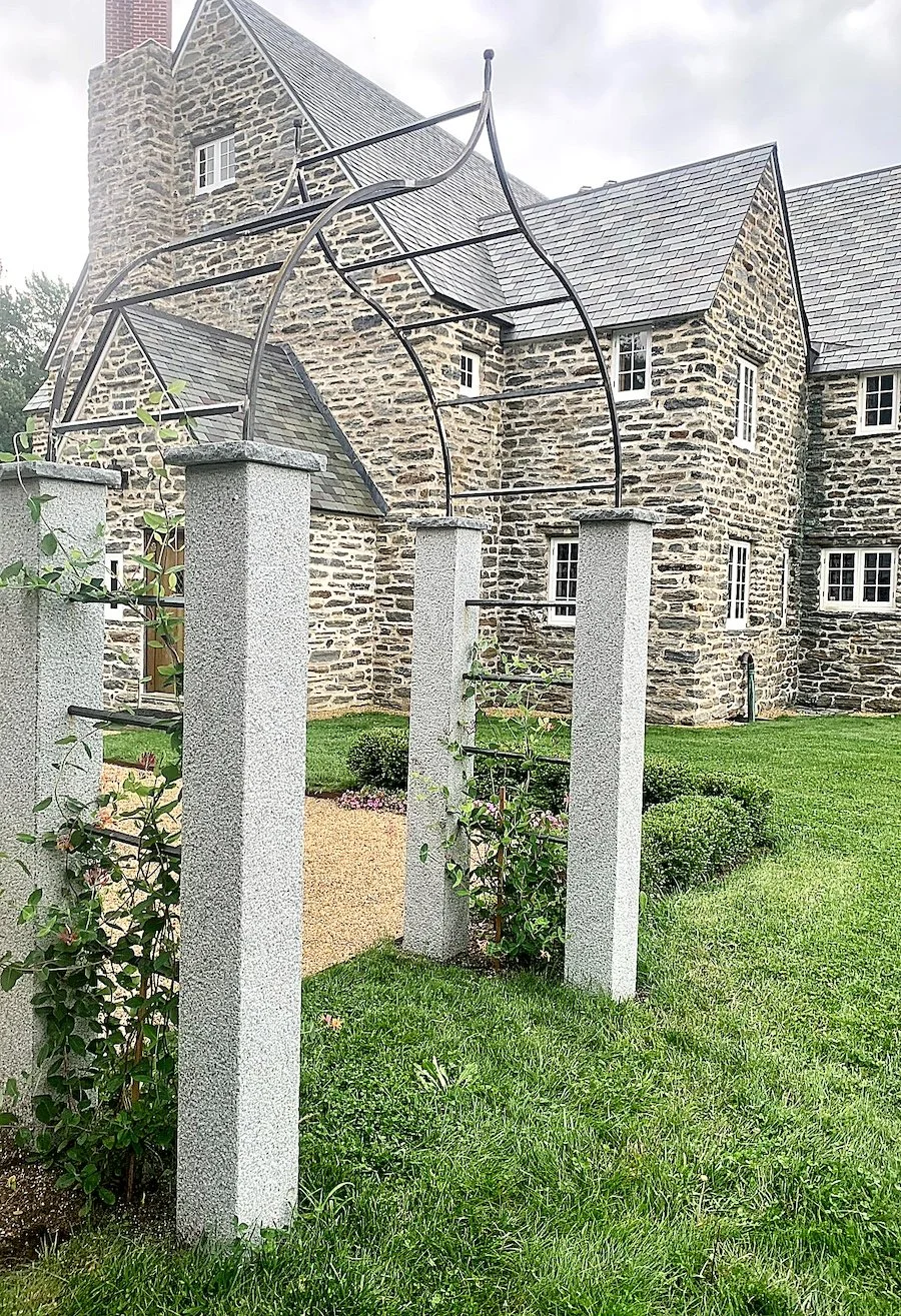 Stone house with grey shingle roof and small white window panes, surrounded by a green lawn and shrubs, with a metal garden archway and concrete pillars in the foreground.