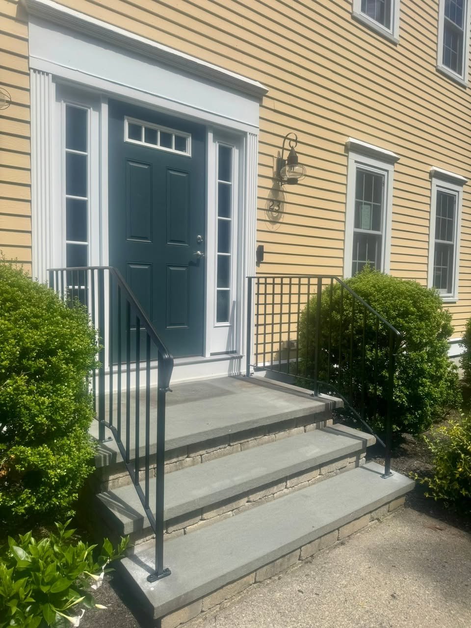 Front porch with four concrete steps, black metal railings, a dark blue front door with sidelights, porch light, and yellow siding house.