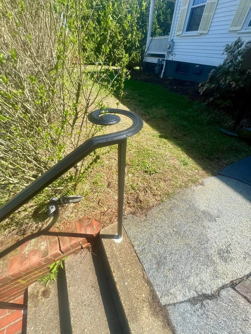 View of a sidewalk and front yard with a metal handrail, brick and concrete steps, shrubs, grass, and a white house in the background.