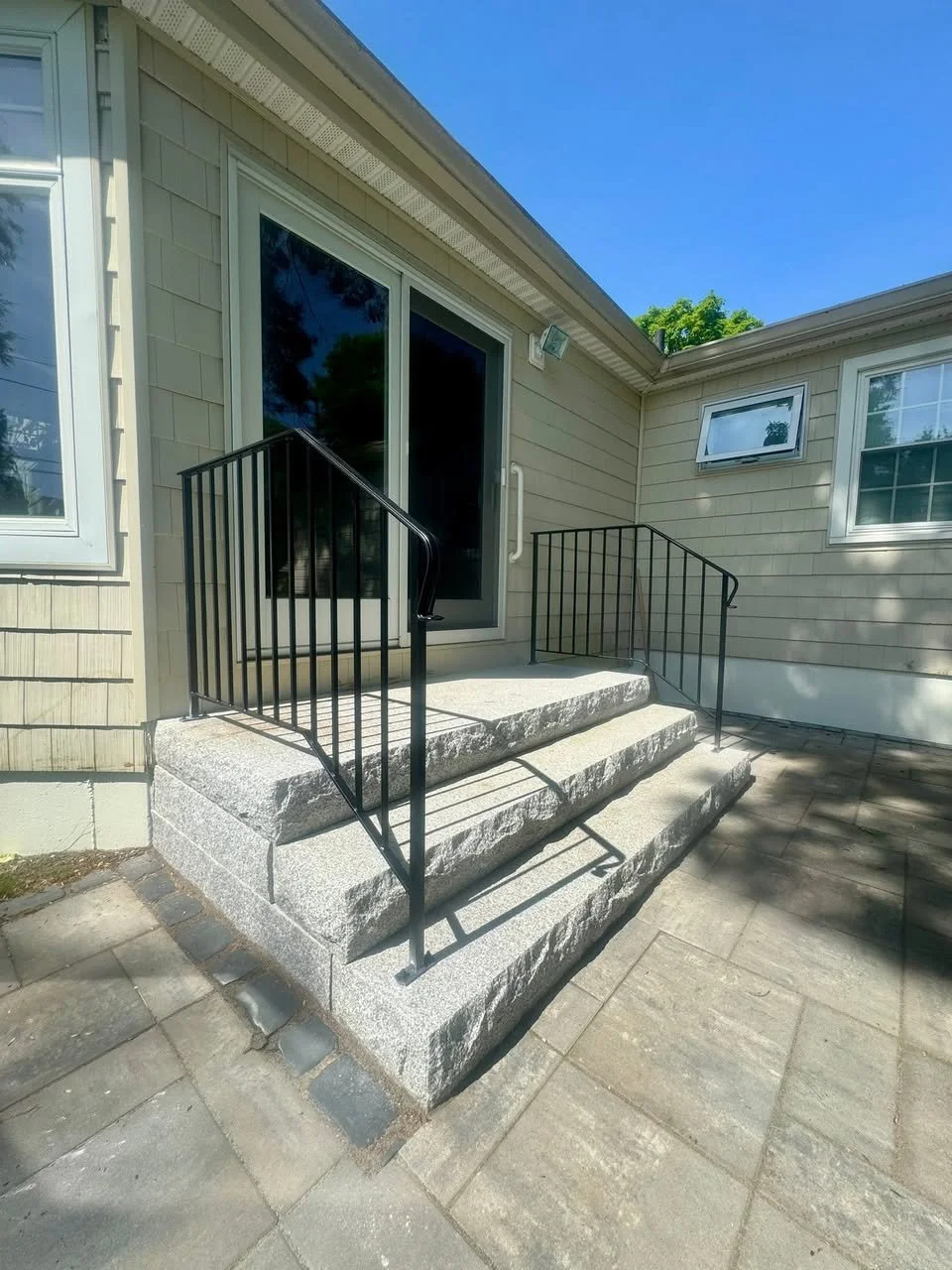 Exterior view of a house with gray siding and a concrete entrance with three steps. The steps have black metal railings on either side and lead to a sliding glass door. There are two windows, one small window to the right of the door and a larger win
