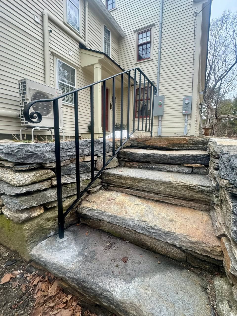 Stone stairs leading to a house with a black metal railing on one side, beige siding, and electrical meters on the exterior wall.