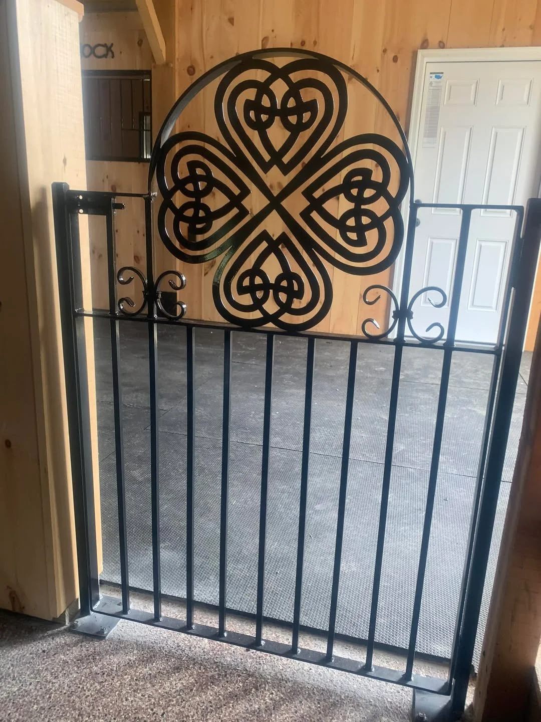 Black metal garden gate featuring a decorative four-leaf clover pattern at the top, installed on a wooden structure inside a room with white closet door in the background.