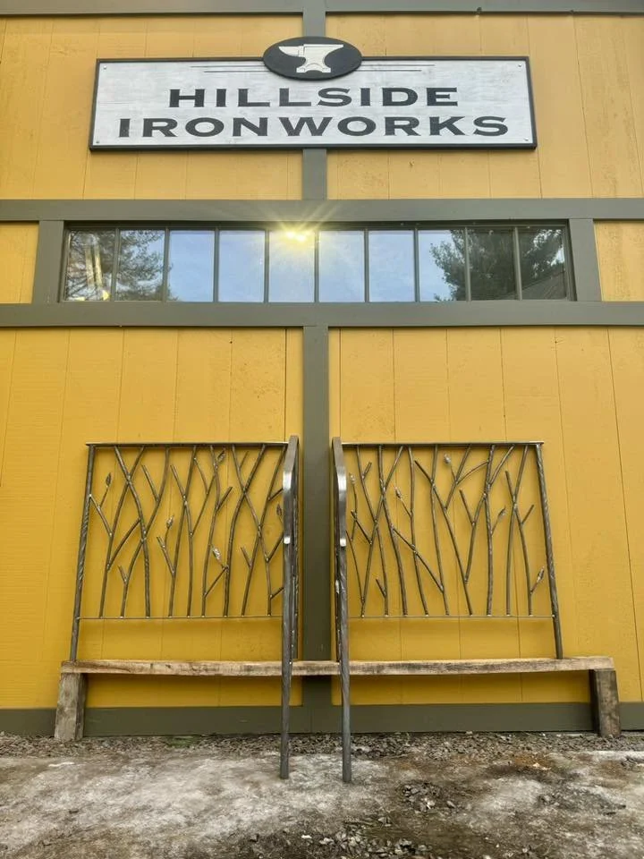 Yellow building with a sign that reads 'Hillside Ironworks' and a logo of a hammer above it, metal gate in front of the building with a design resembling tree branches.