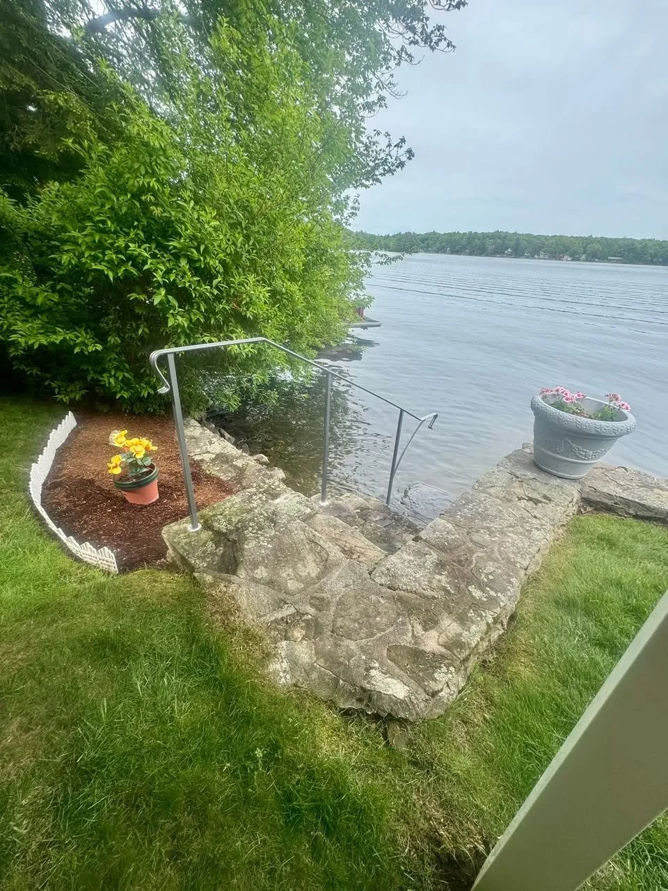 A lakeside scene with a stone patio, metal handrail, and potted flowers overlooking the water, surrounded by greenery.