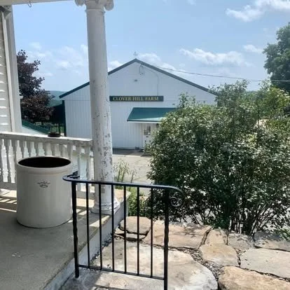 View from a porch with a black railing, a large white trash bin, and a small tree in front of a white farm building with a green sign that says 'Clover Hill Farms'.
