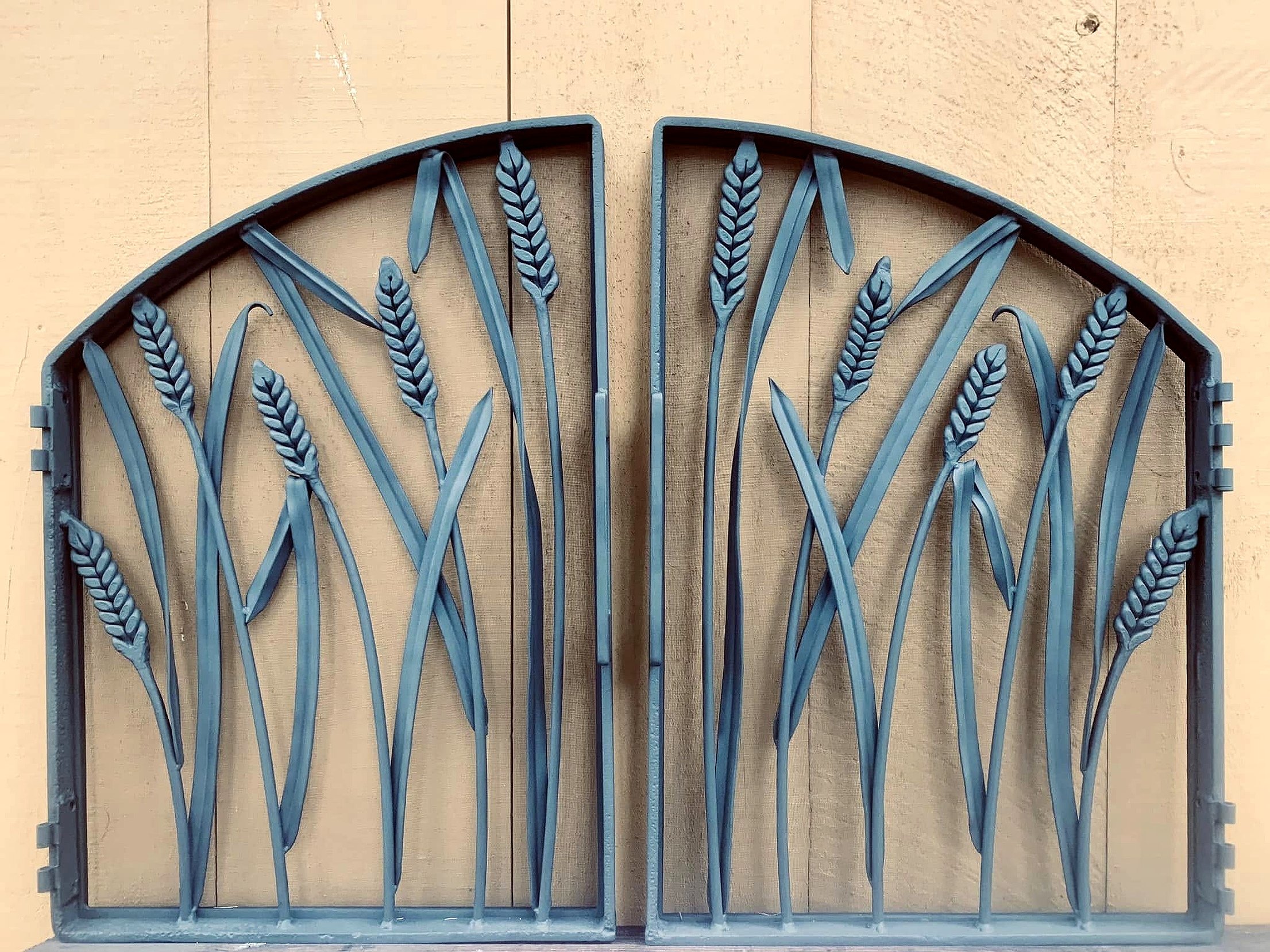 Decorative metal gate with wheat stalks design in front of a beige wall.
