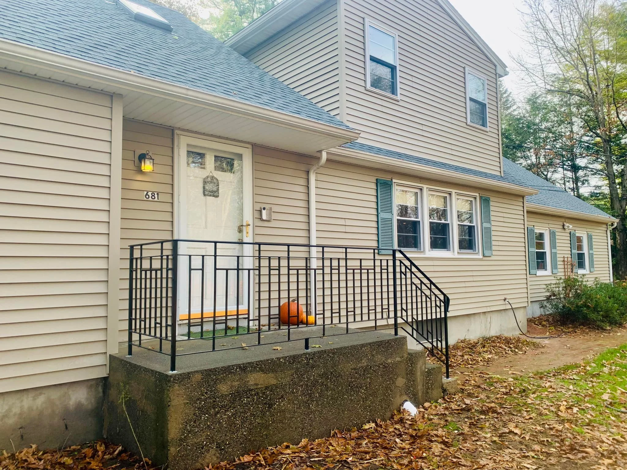 The front of a two-story house with beige siding, green shutters, and a blue roof, featuring a small porch with a black metal railing and pumpkins near the door, surrounded by trees and fallen leaves.