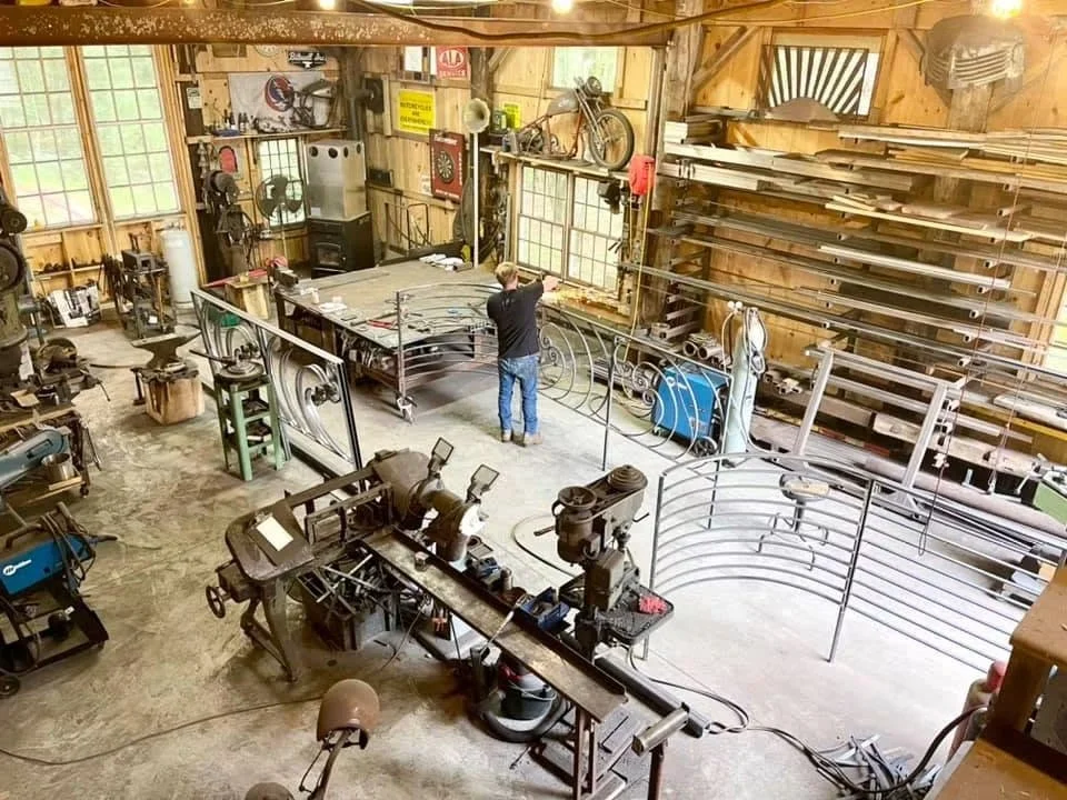 Inside a woodworking shop with a person working on a metal railing or fence. The workshop contains various tools, equipment, and materials, with wooden shelves stocked with lumber and large windows letting in natural light.
