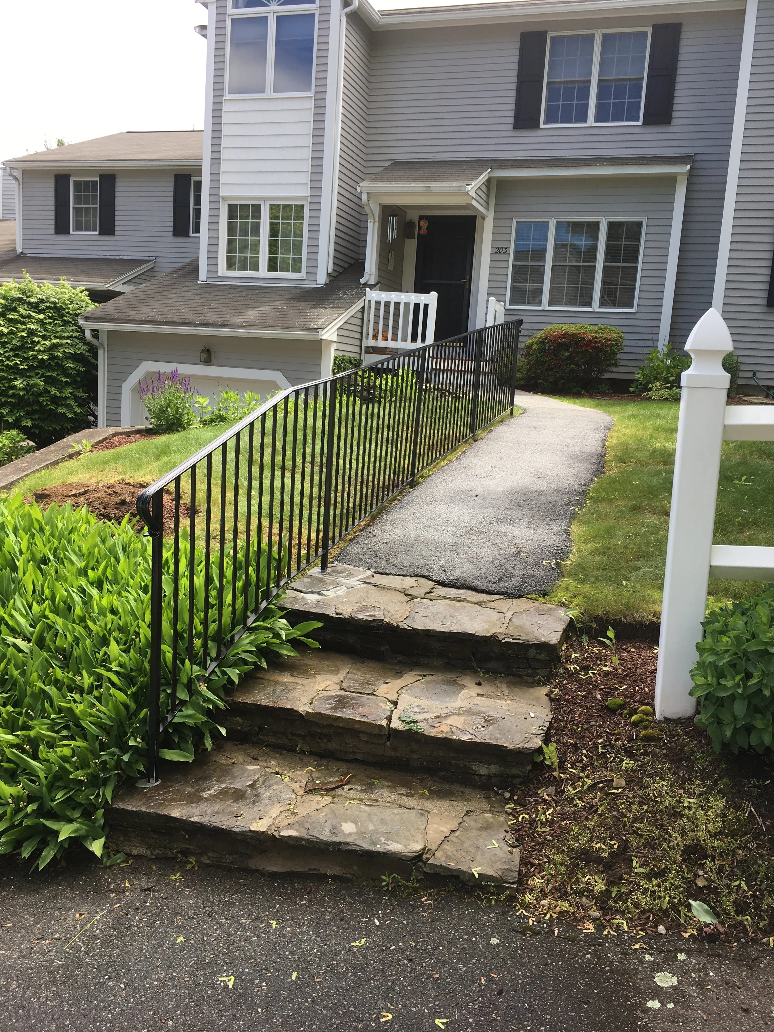 The image shows a multi-story house with gray siding, black shutters, and large windows. A small porch with a black door is accessible via a curved paved walkway with stone stairs. There is greenery, plants, and bushes around the house, with flower b