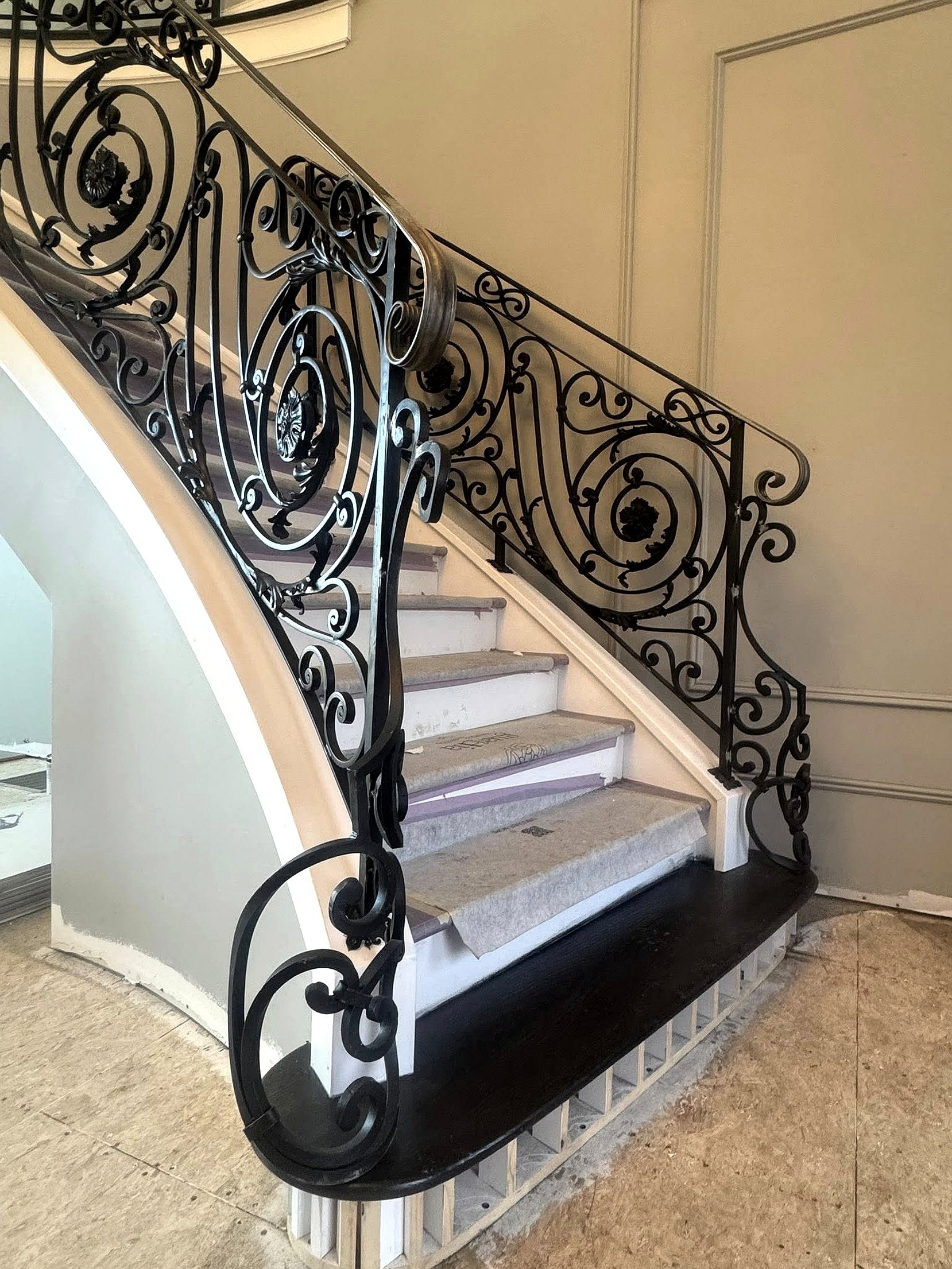 Wooden staircase with decorative wrought iron railing leading to an upper floor, beside a beige wall with decorative trim, in a building interior.