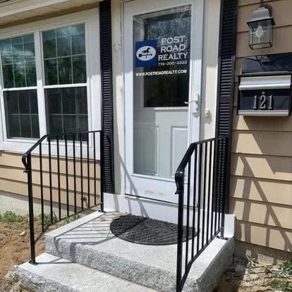 Front door with a sign that reads Post Road Realty, black metal handrails, grey concrete steps, black doormat, and a mailbox with the number 121 in black.